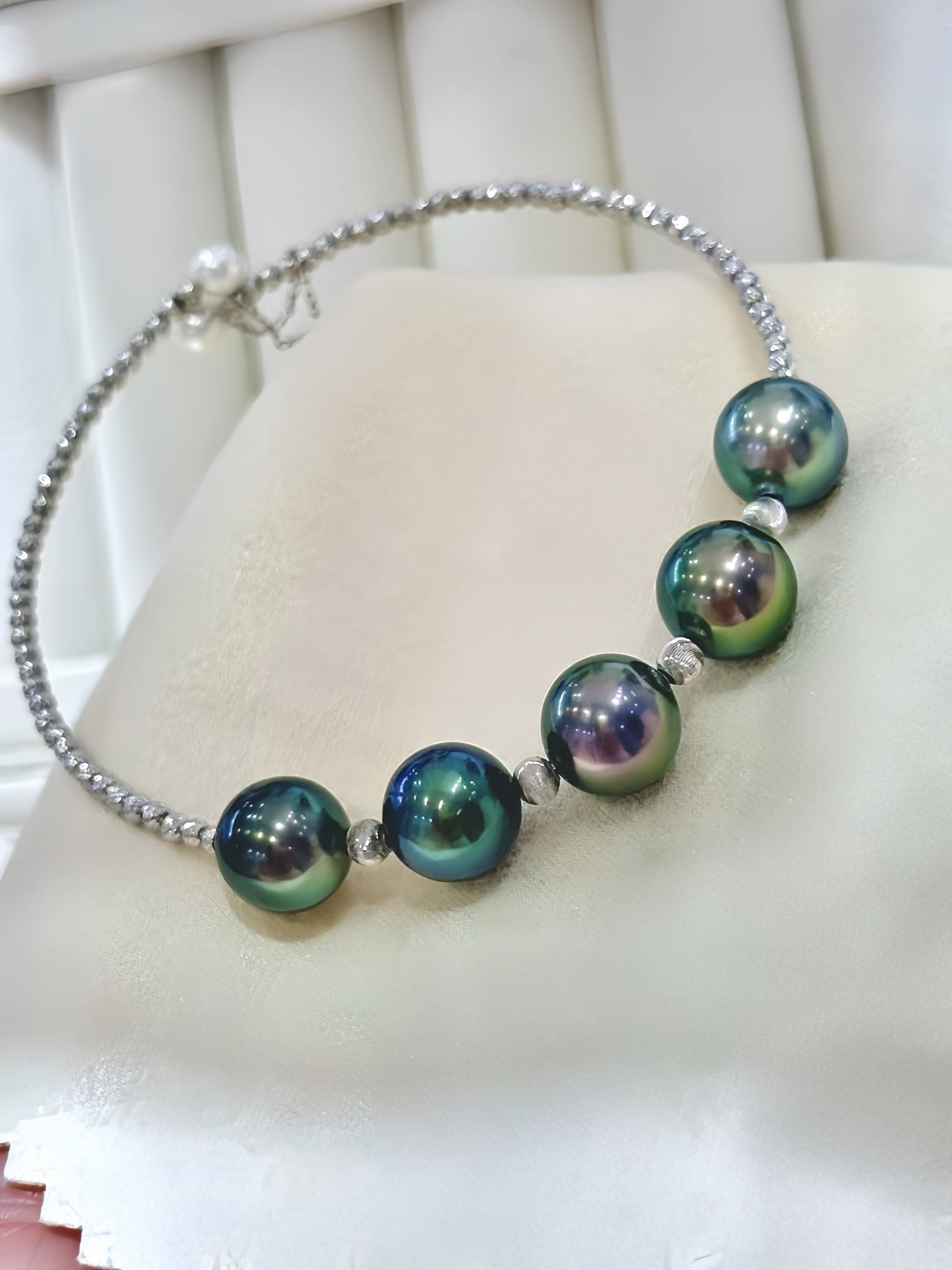A beaded silver chain necklace featuring five large, iridescent green and blue pearls separated by small silver beads, displayed on a cream-colored jewelry stand.