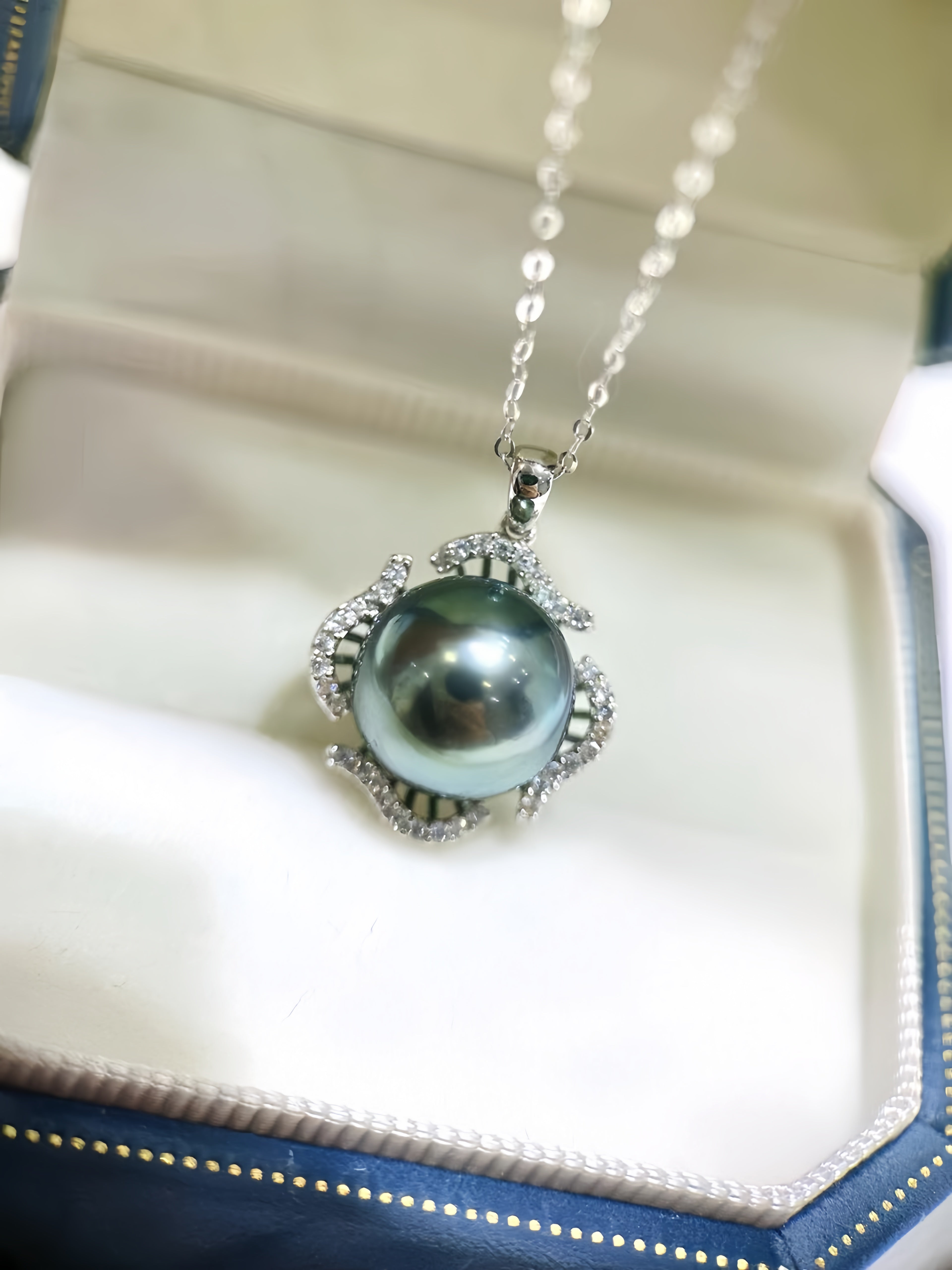 A silver pendant necklace featuring a large, round, dark green-gray pearl surrounded by small clear stones, resting inside an open cream-colored jewelry box.