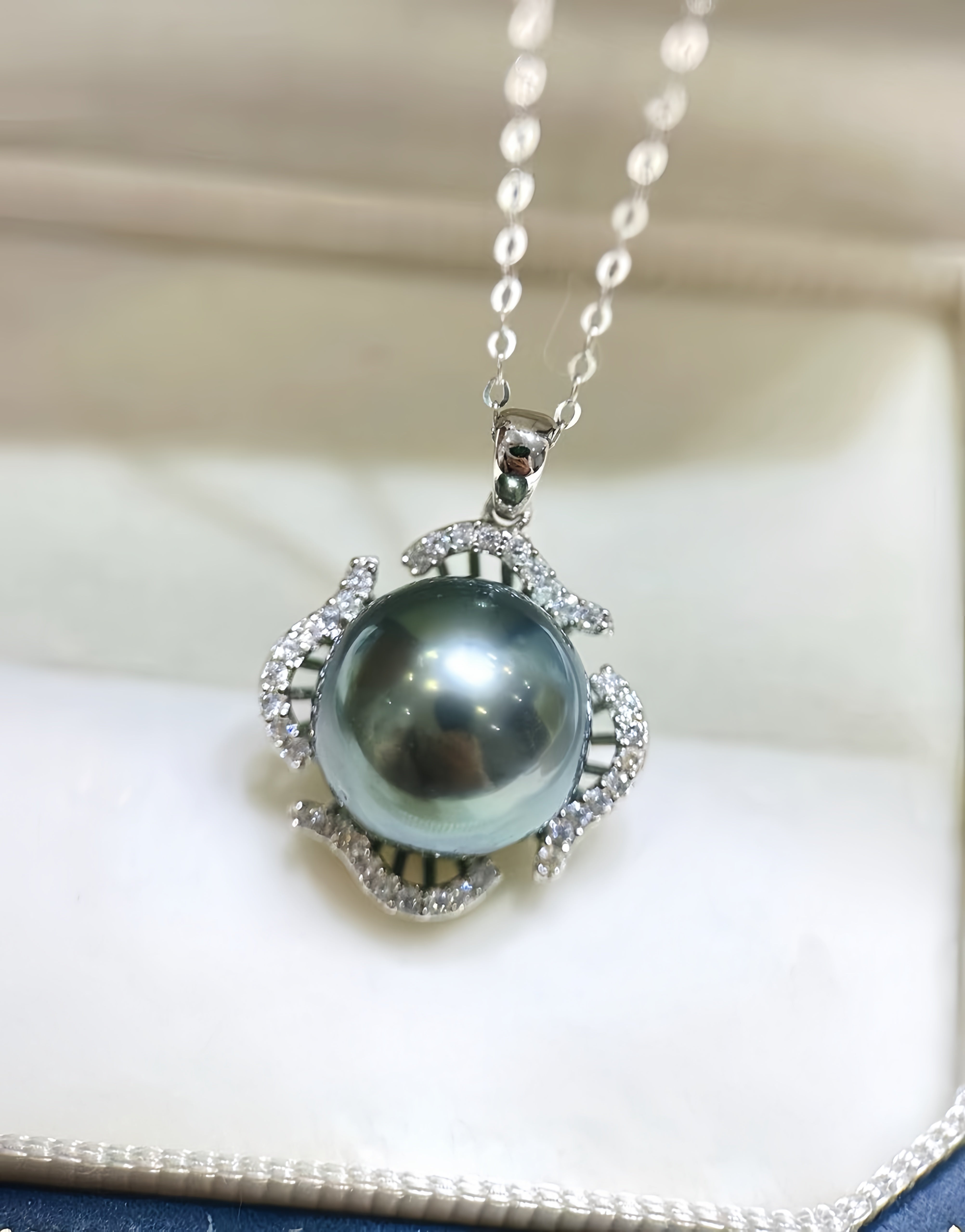 A silver pendant necklace featuring a large, lustrous dark gray pearl surrounded by small clear stones, resting on a white jewelry box.