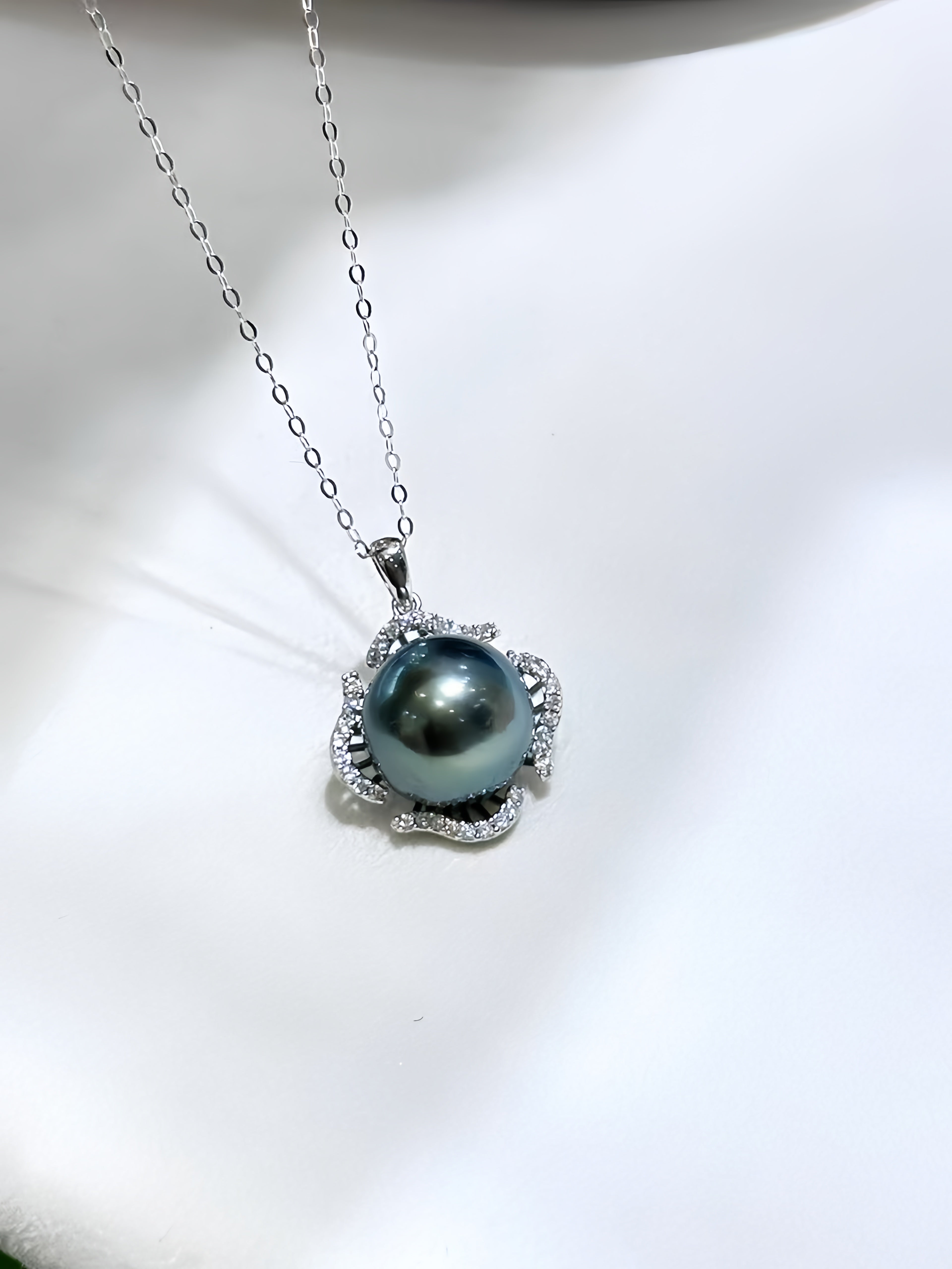 A silver pendant necklace featuring a round, dark blue-gray pearl surrounded by small clear stones, displayed on a white satin surface.