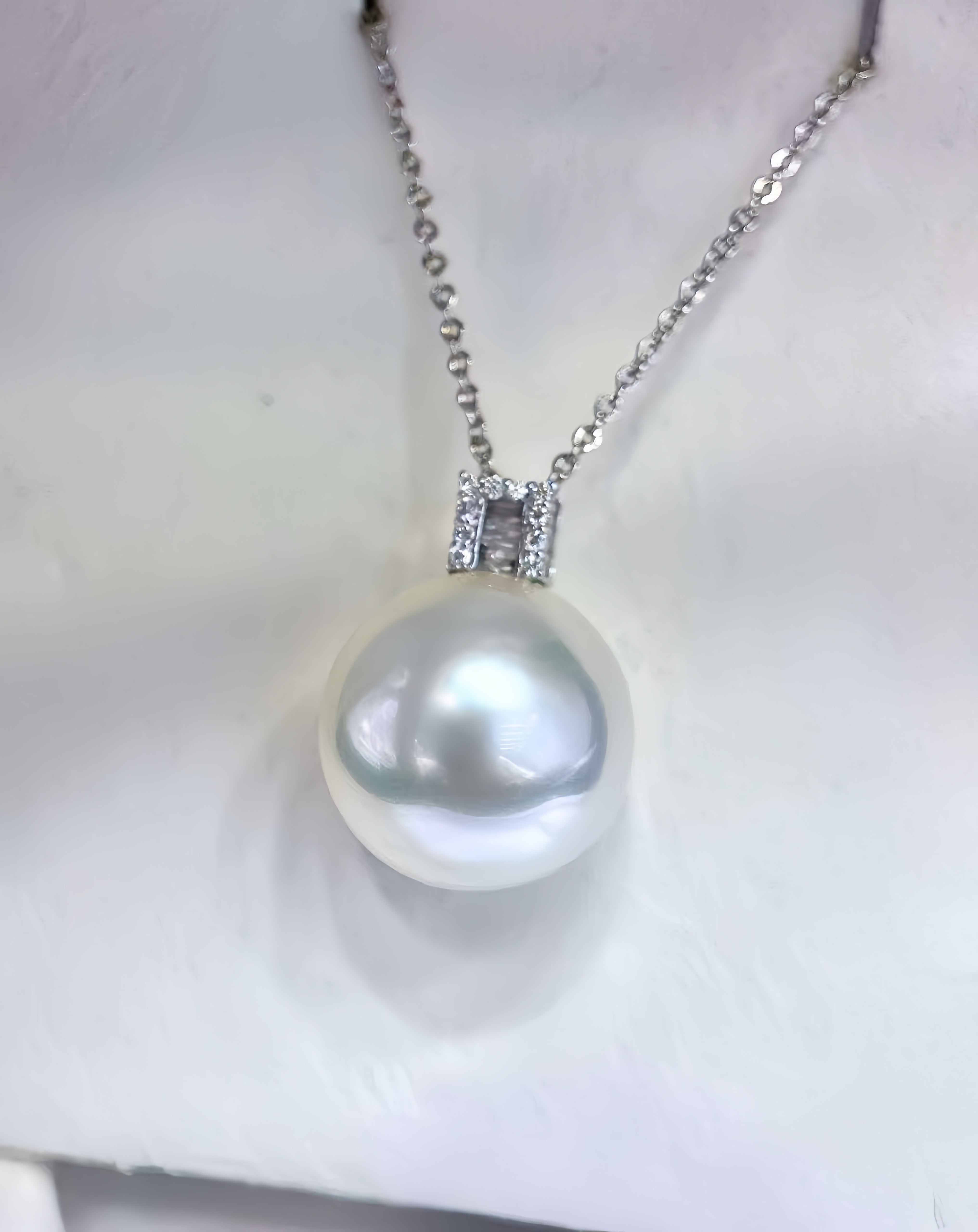 A silver chain necklace featuring a large, lustrous white pearl pendant set in a silver bezel with small clear stones, displayed on a white textured surface.