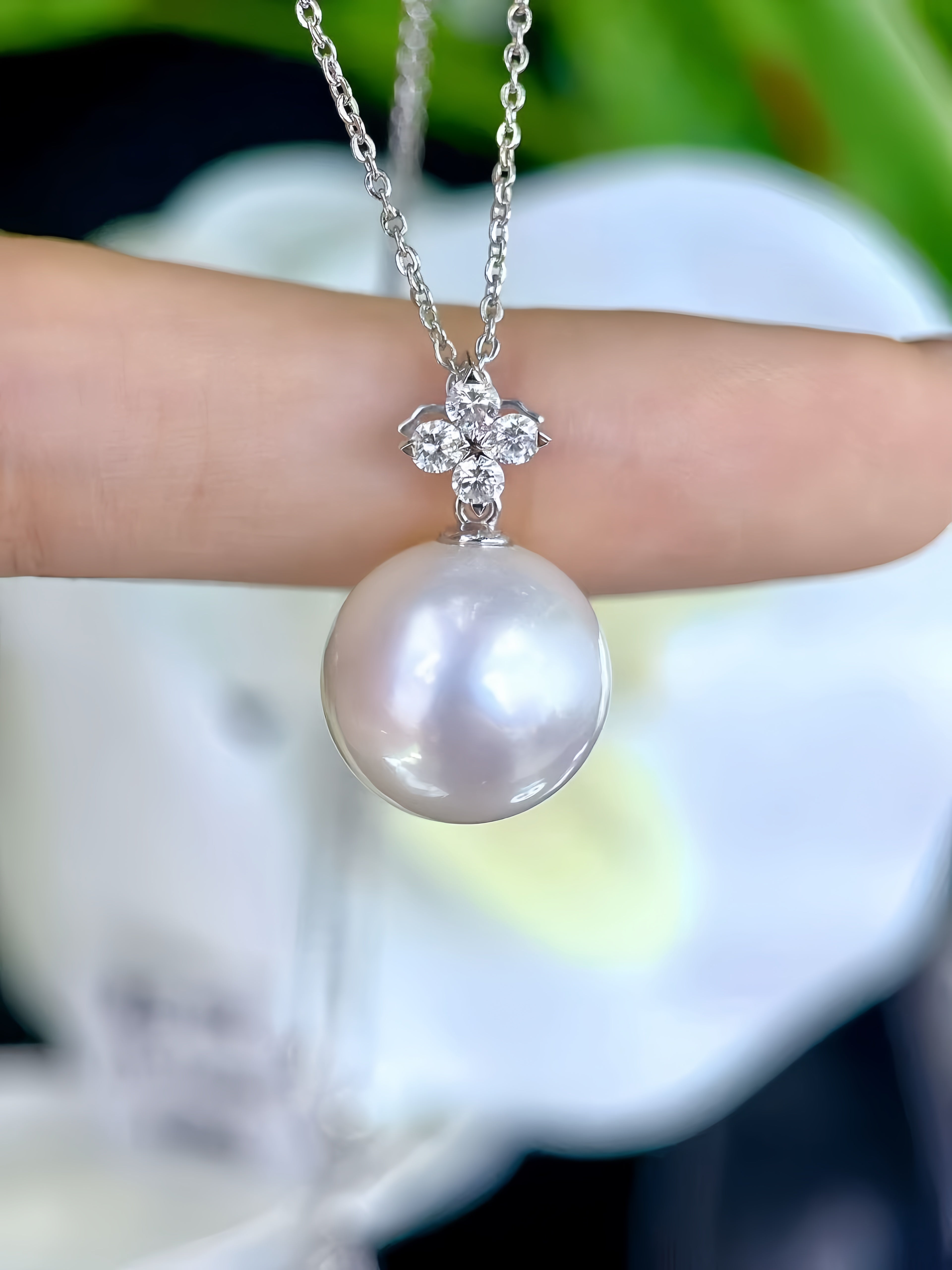 A silver chain necklace with a large, round white pearl pendant and a small floral cluster of clear stones at the top, shown resting on a person's finger.
