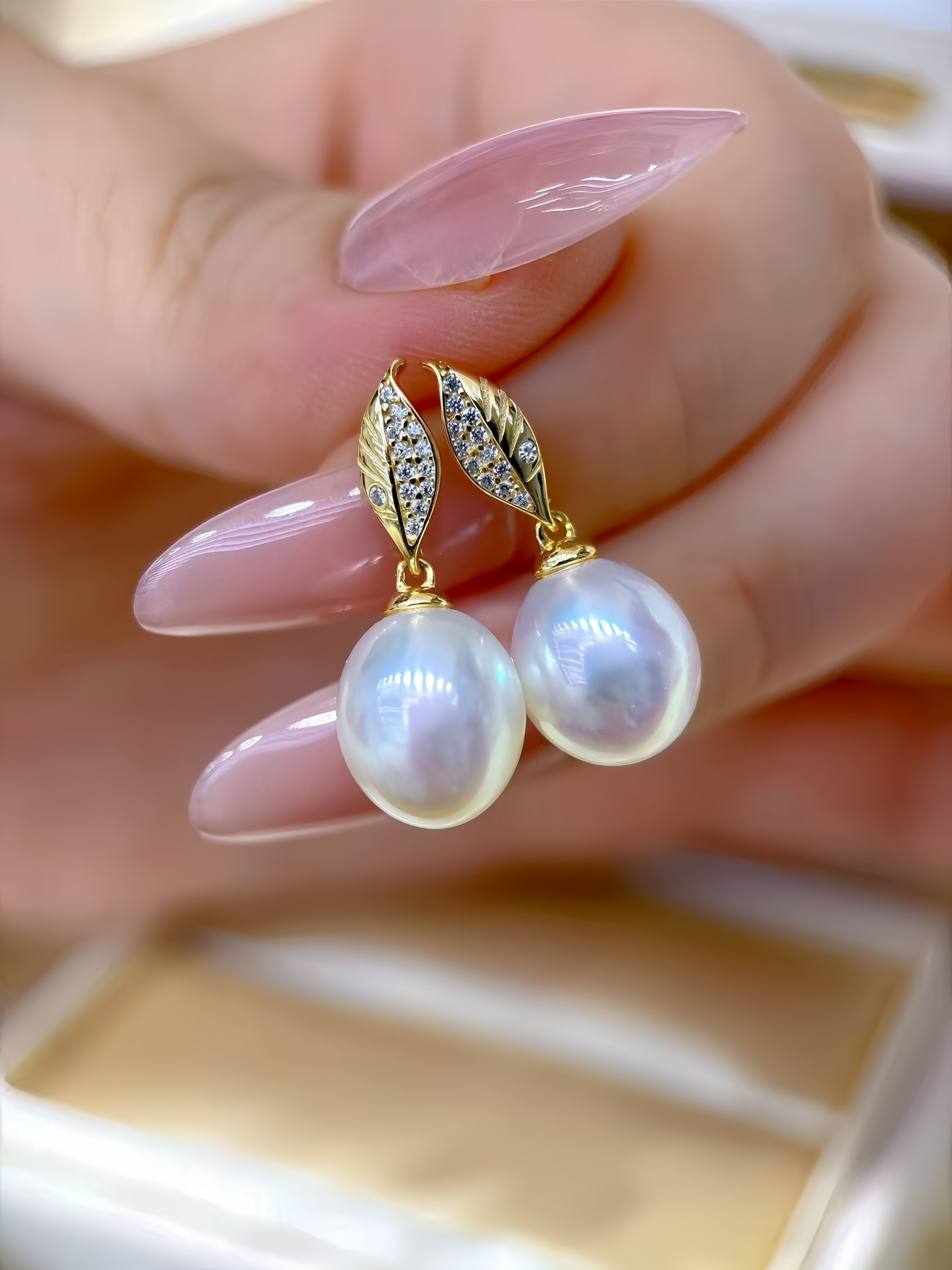 A close-up of a hand with long pink manicured nails holding a pair of gold leaf-shaped drop earrings with small clear stones and large white pearl-like drops.