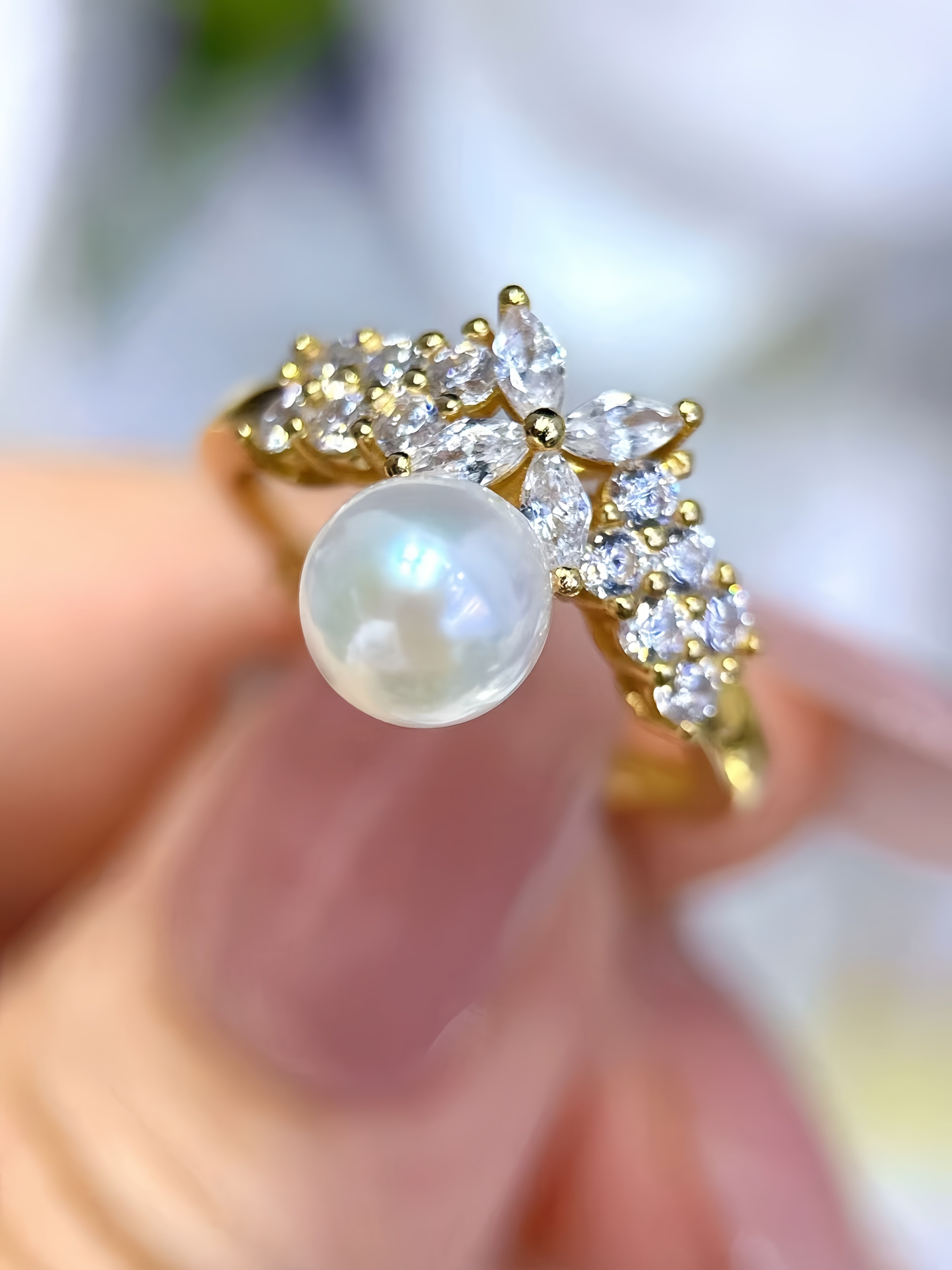 A gold ring with a large round white pearl and small clear stones, held between fingers against a soft blurred background.