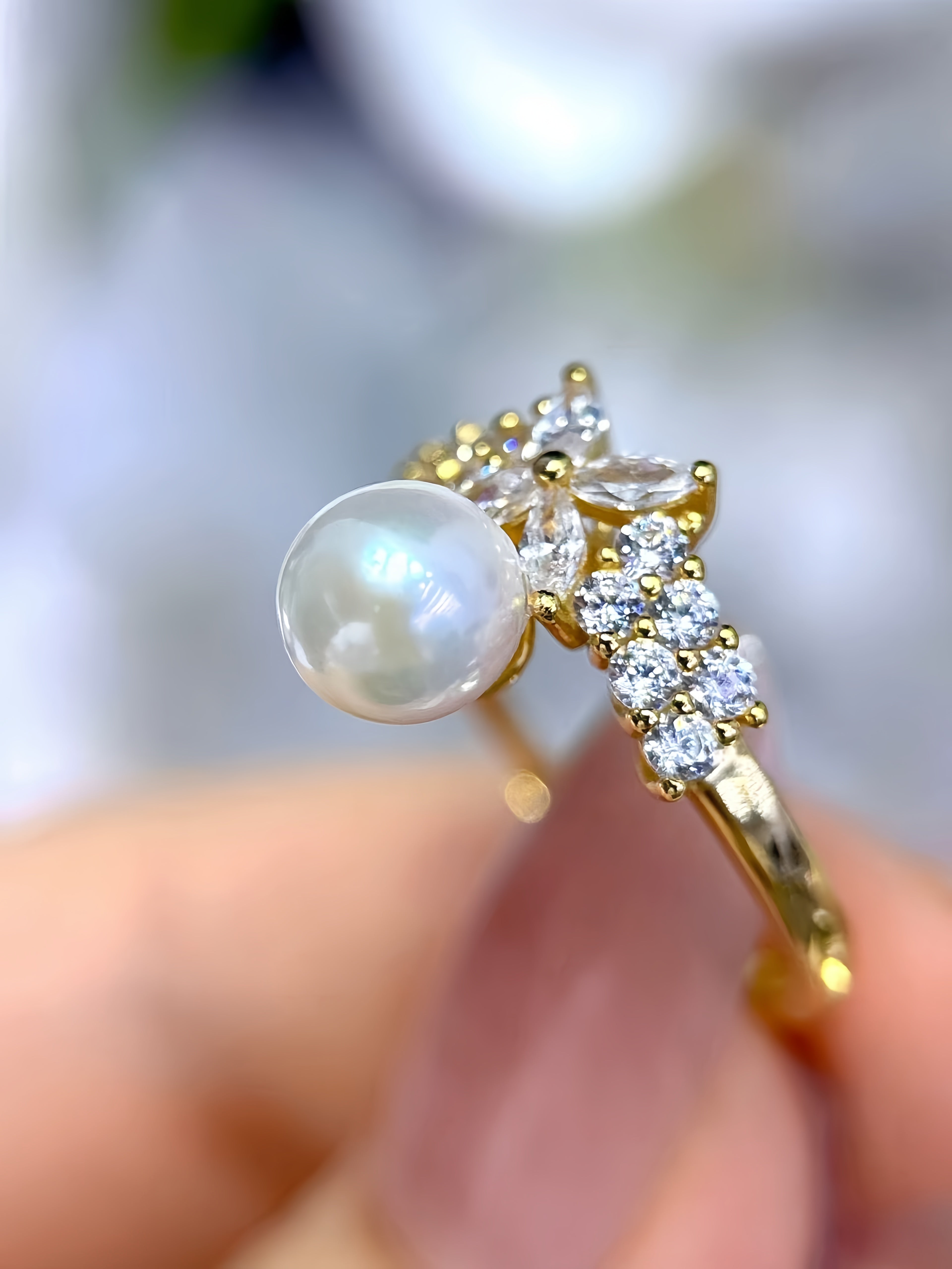 A gold ring with a large round white pearl and small clear stones, held between fingers against a soft blurred background.