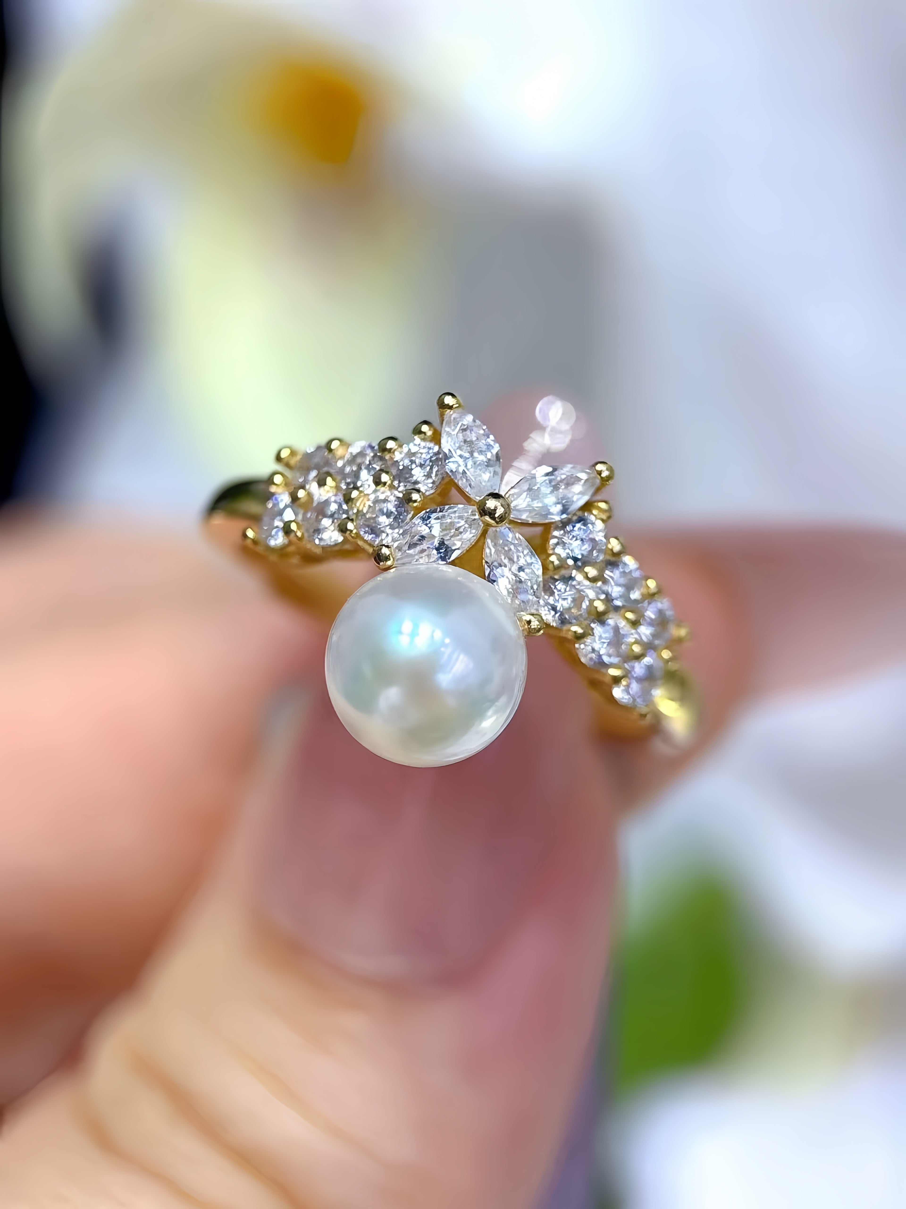 A gold ring with a large white pearl and clear marquise and round-cut stones, held between fingers against a soft, blurred white and green background.