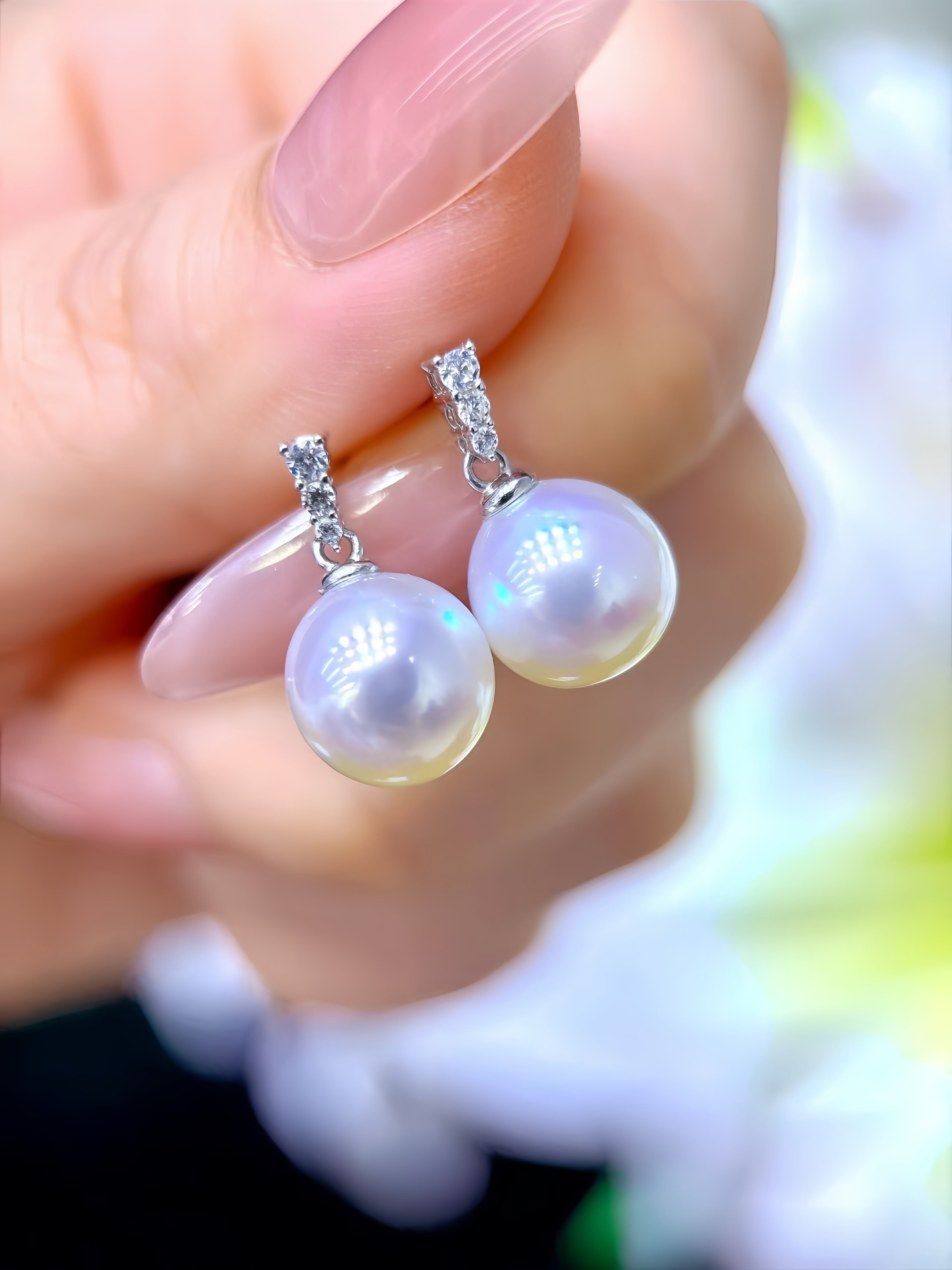 A close-up of a hand holding a pair of silver drop earrings with large lustrous pearl-like beads and small clear stones at the top, against a softly blurred light background.