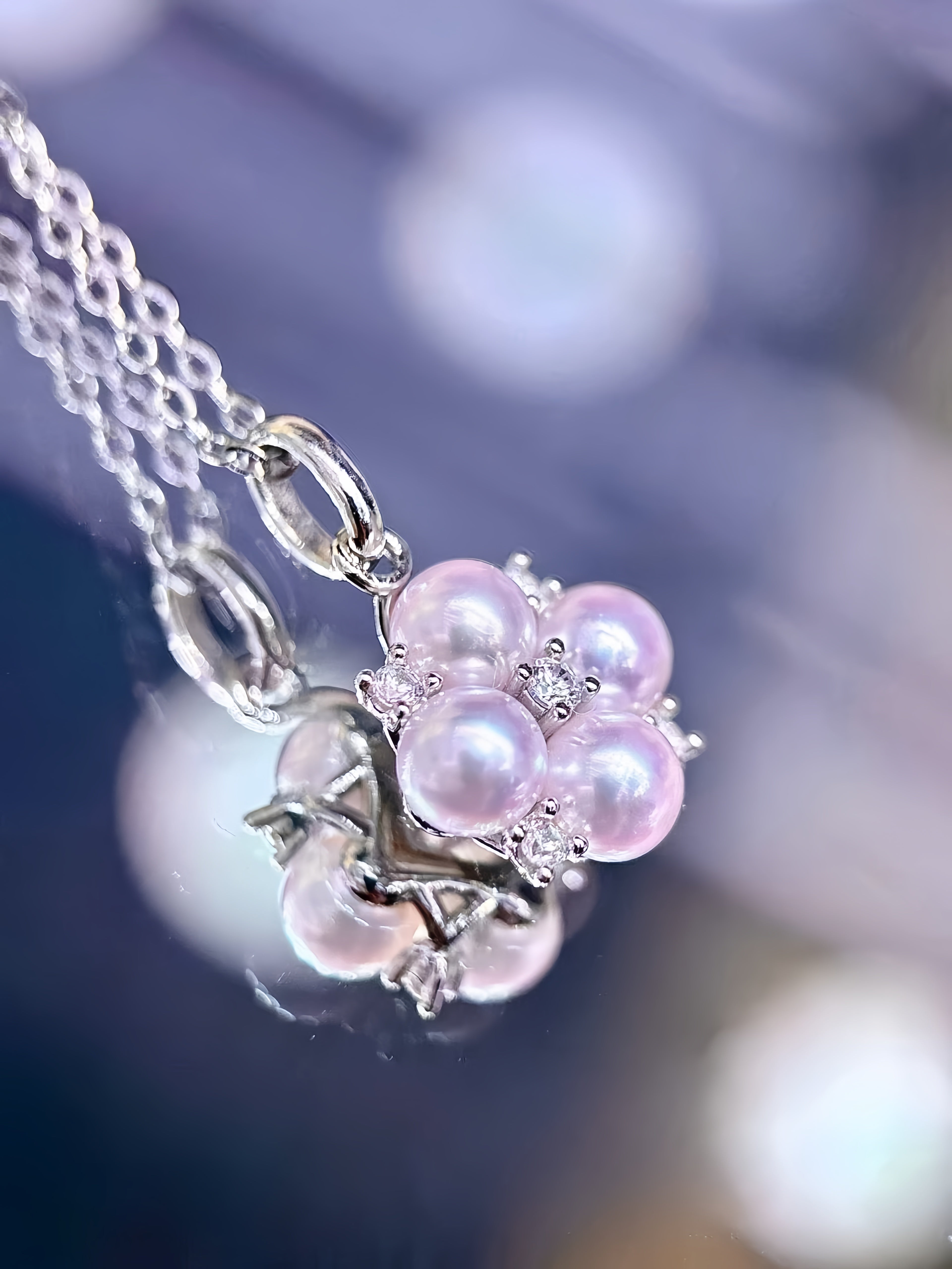 A silver pendant necklace featuring a cluster of iridescent light pink freshwater pearls and small clear crystal accents, shown close-up against a soft, out-of-focus blue and purple background.