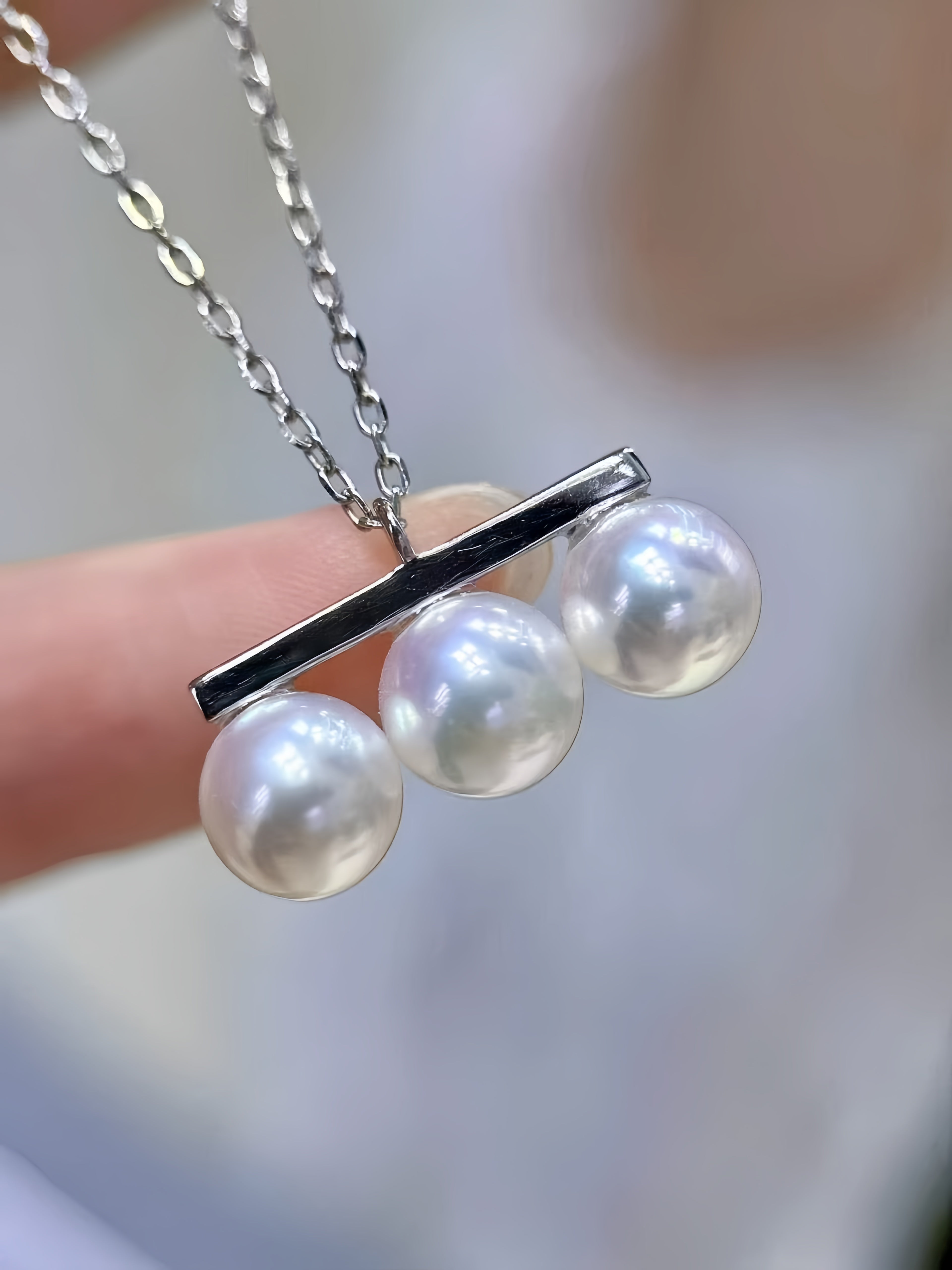 A close-up of a silver necklace with a bar pendant holding three round white pearls, held by a hand against a soft blurred background.