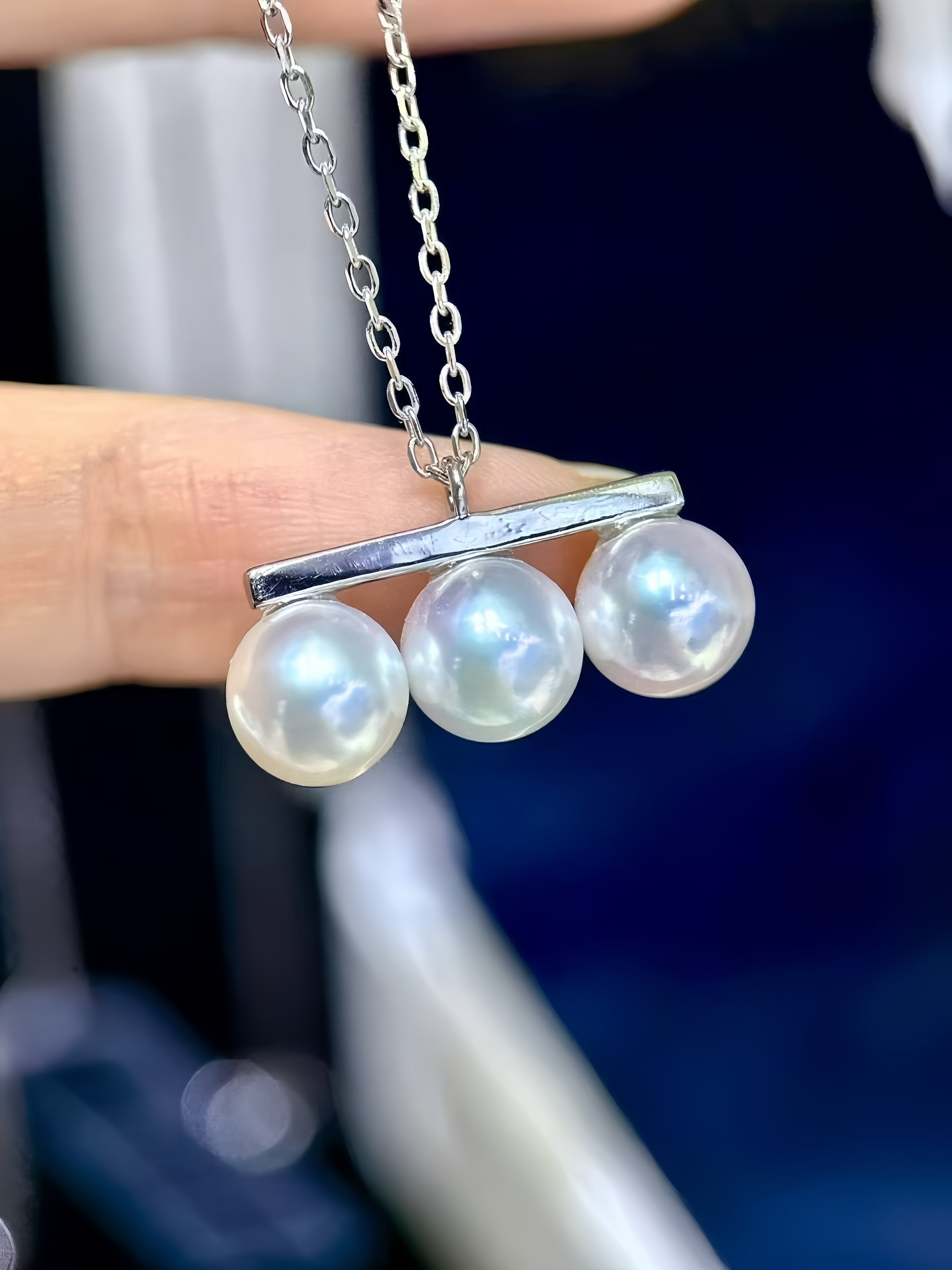 A close-up of a silver necklace with three large, lustrous white freshwater pearls suspended from a horizontal bar, held by a person's fingers against a dark blue and white blurred background.