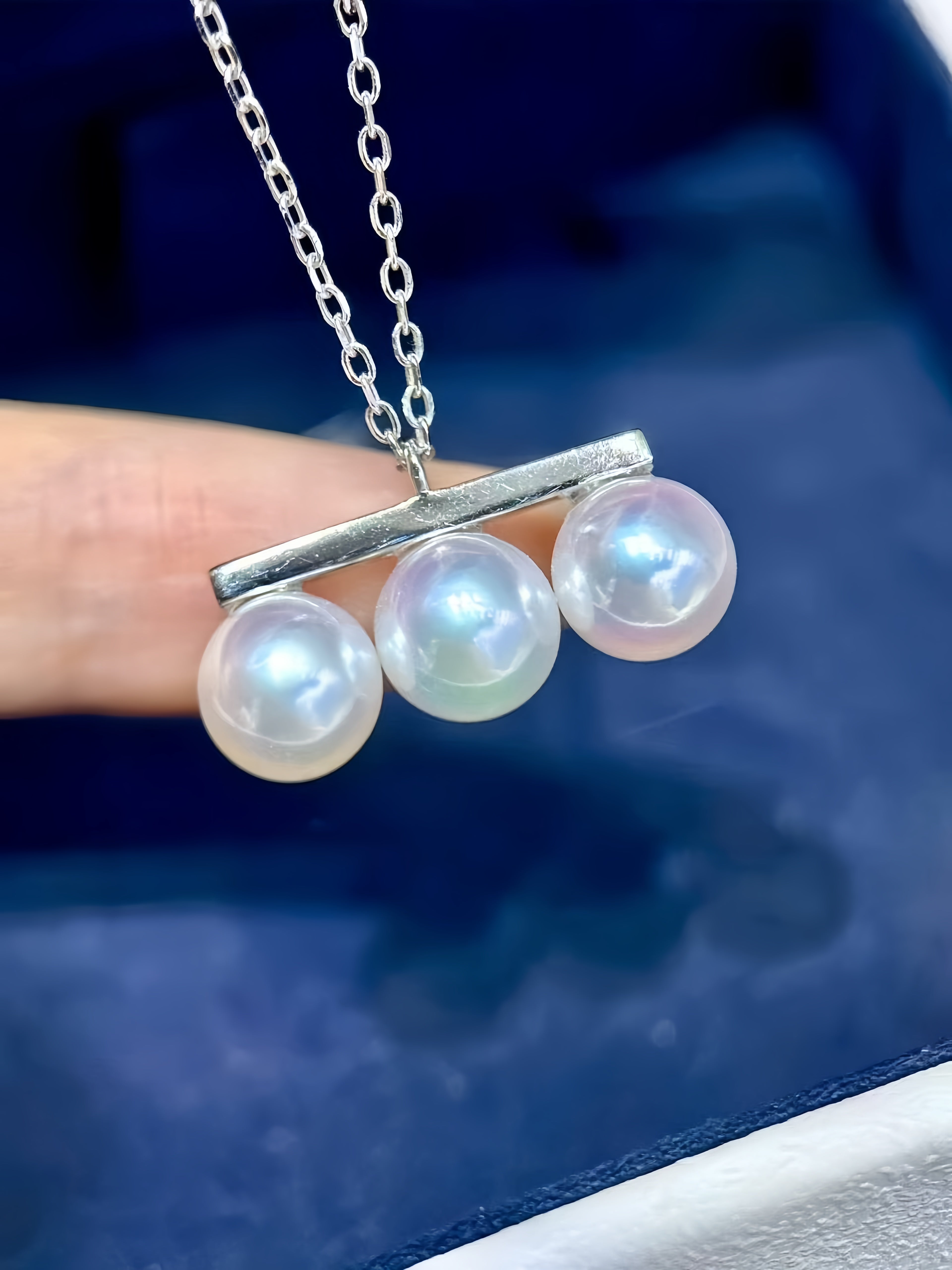 A close-up of a silver necklace with three round iridescent white pearls suspended from a horizontal bar, held by a hand against a dark blue background.
