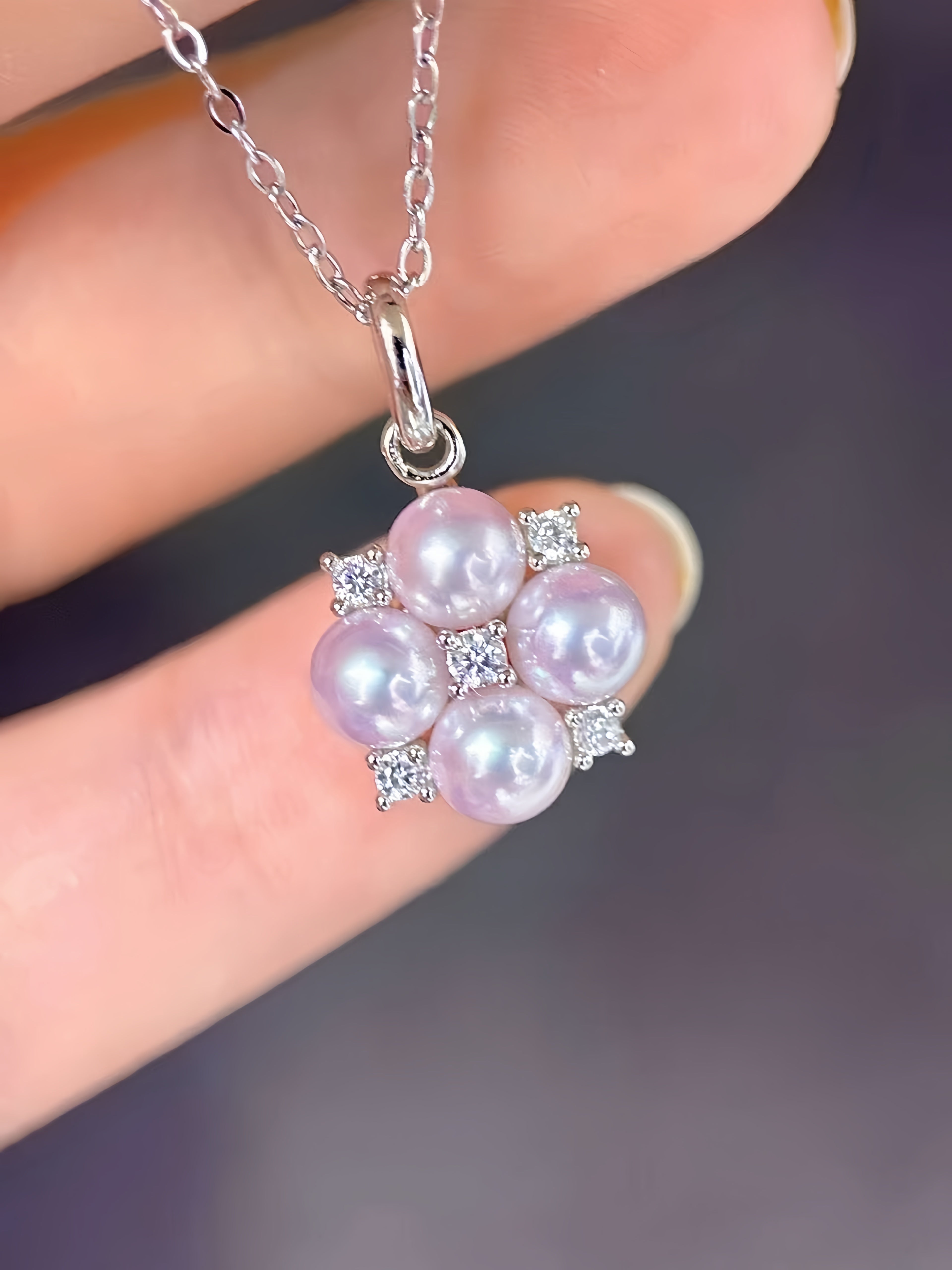 A close-up of a hand holding a silver pendant necklace featuring four light pink freshwater pearls arranged in a flower shape, accented with small clear cubic zirconia stones.