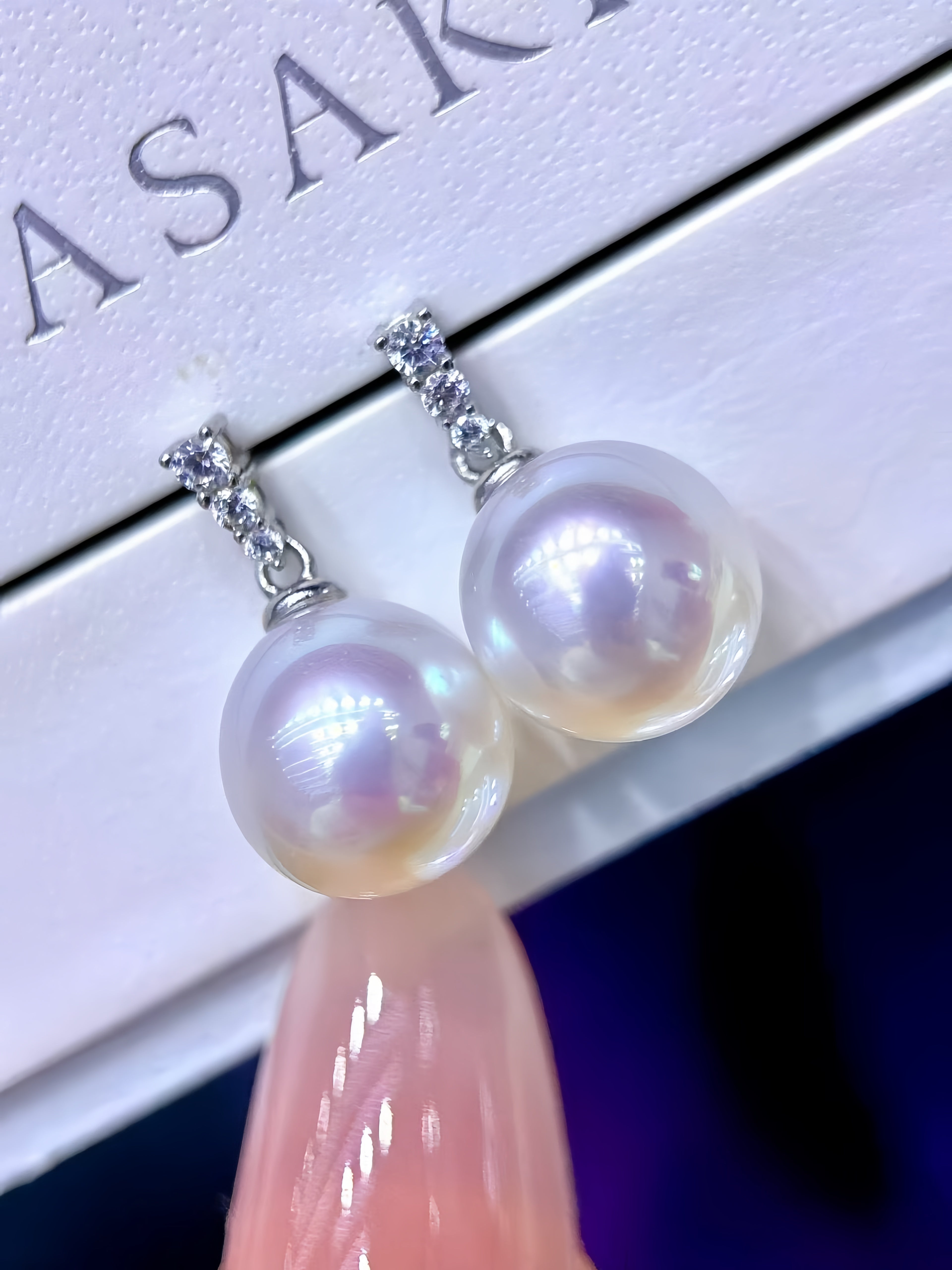A pair of dangling pearl earrings with small clear stone accents at the top, shown resting on a white jewelry box. A finger with a pink manicure is visible at the bottom, and part of the brand name ASAKI is printed on the box.