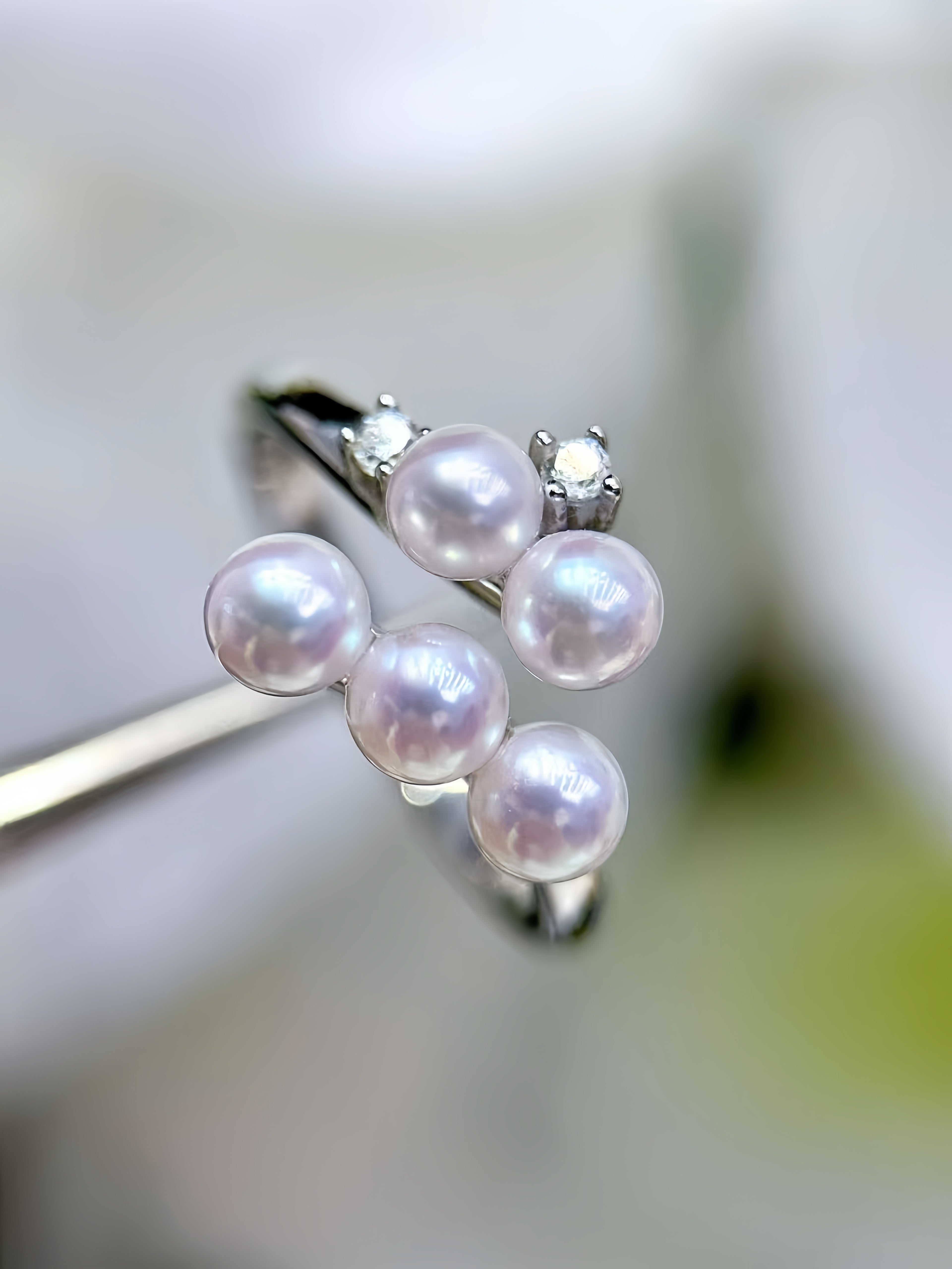A silver ring with five white freshwater pearls clustered on top, held by metal tweezers against a soft, blurred white floral background.