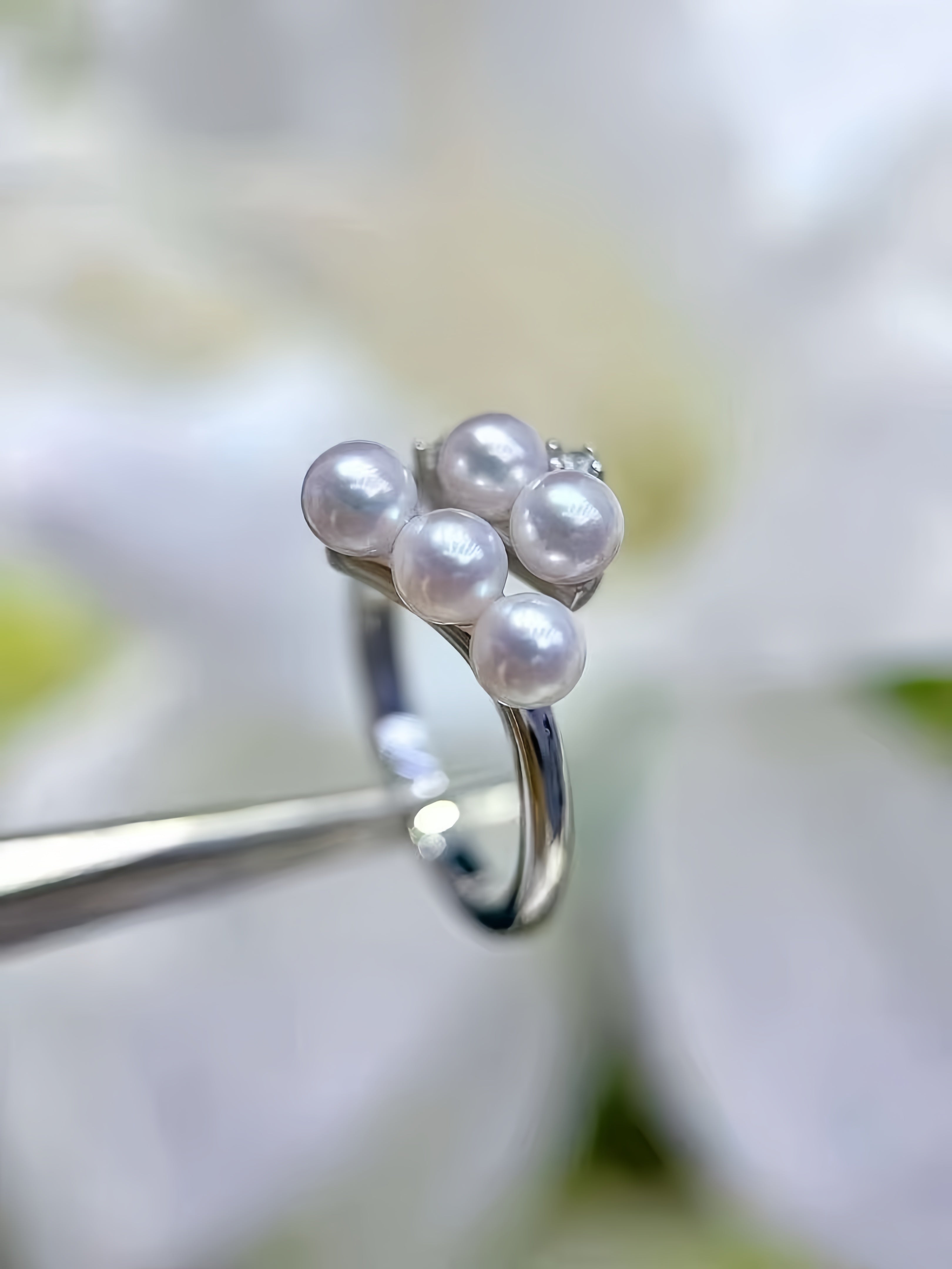 A silver ring with five white freshwater pearls clustered on top, held by metal tweezers against a soft, blurred white floral background.