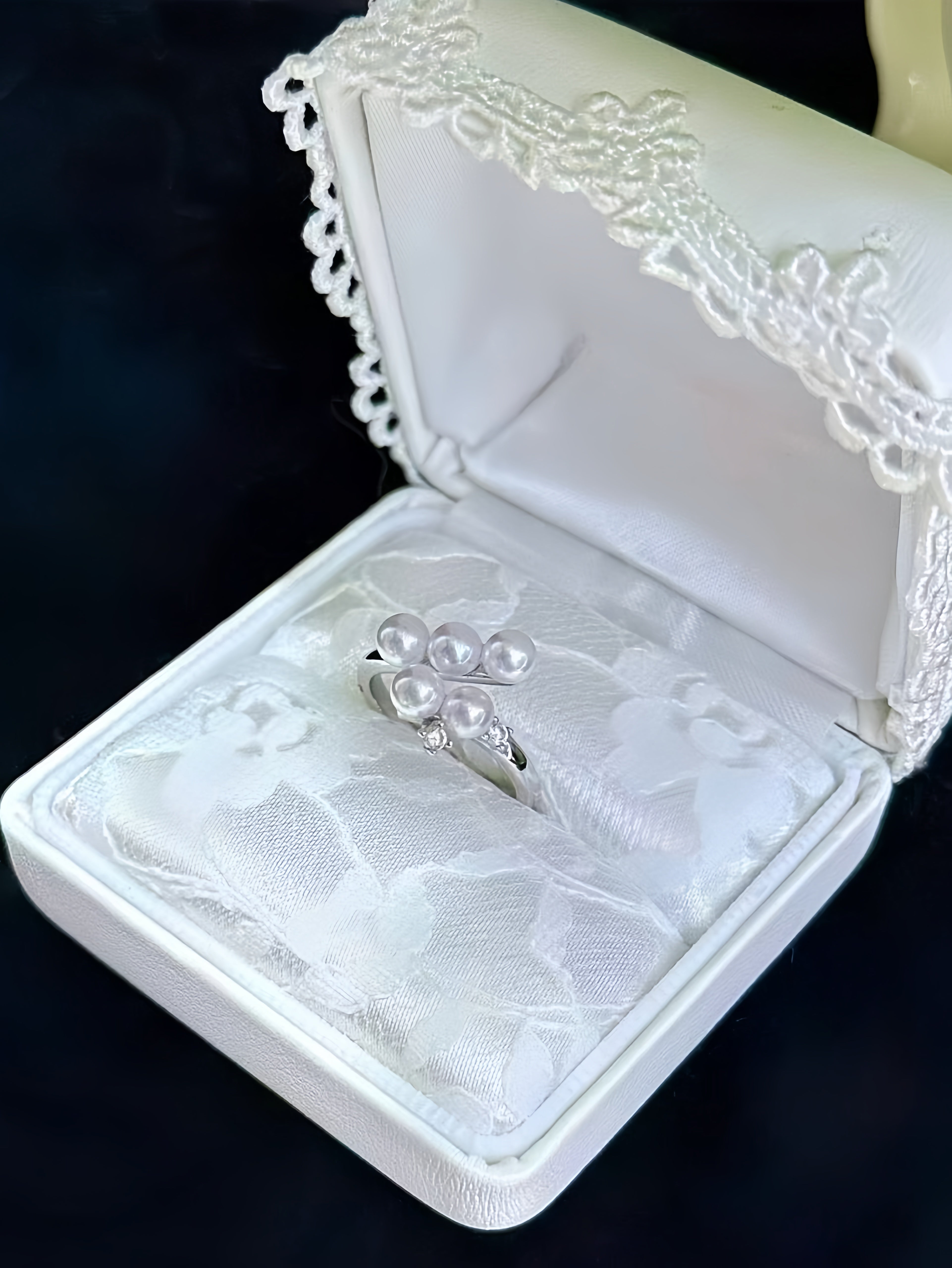 A silver ring with a cluster of white pearls and small clear stones, displayed inside an open white satin ring box with lace trim on a dark background.