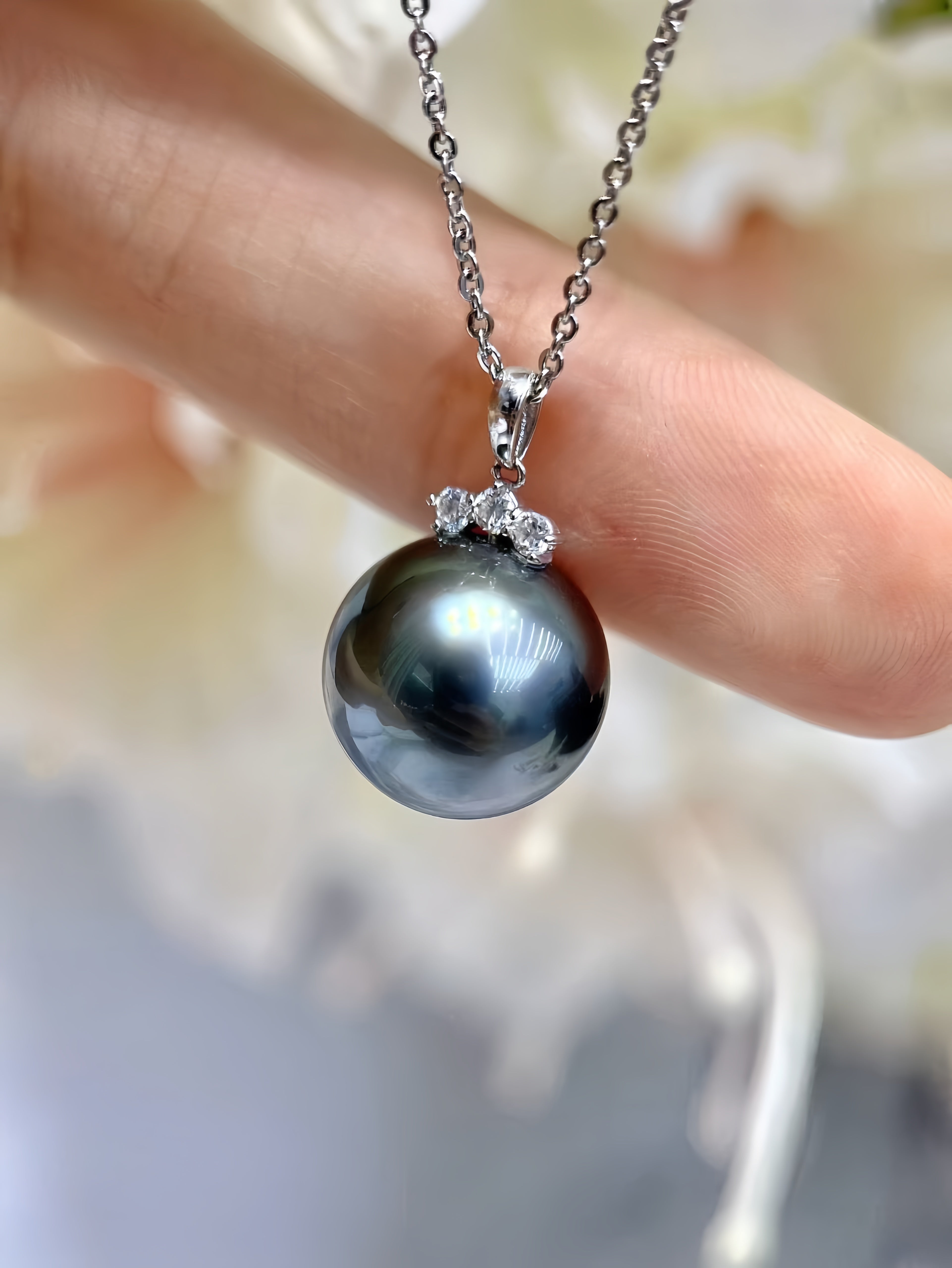 A close-up of a silver chain necklace with a large, round dark gray pearl pendant accented by small clear stones, held against a soft-focus background of skin and white flowers.