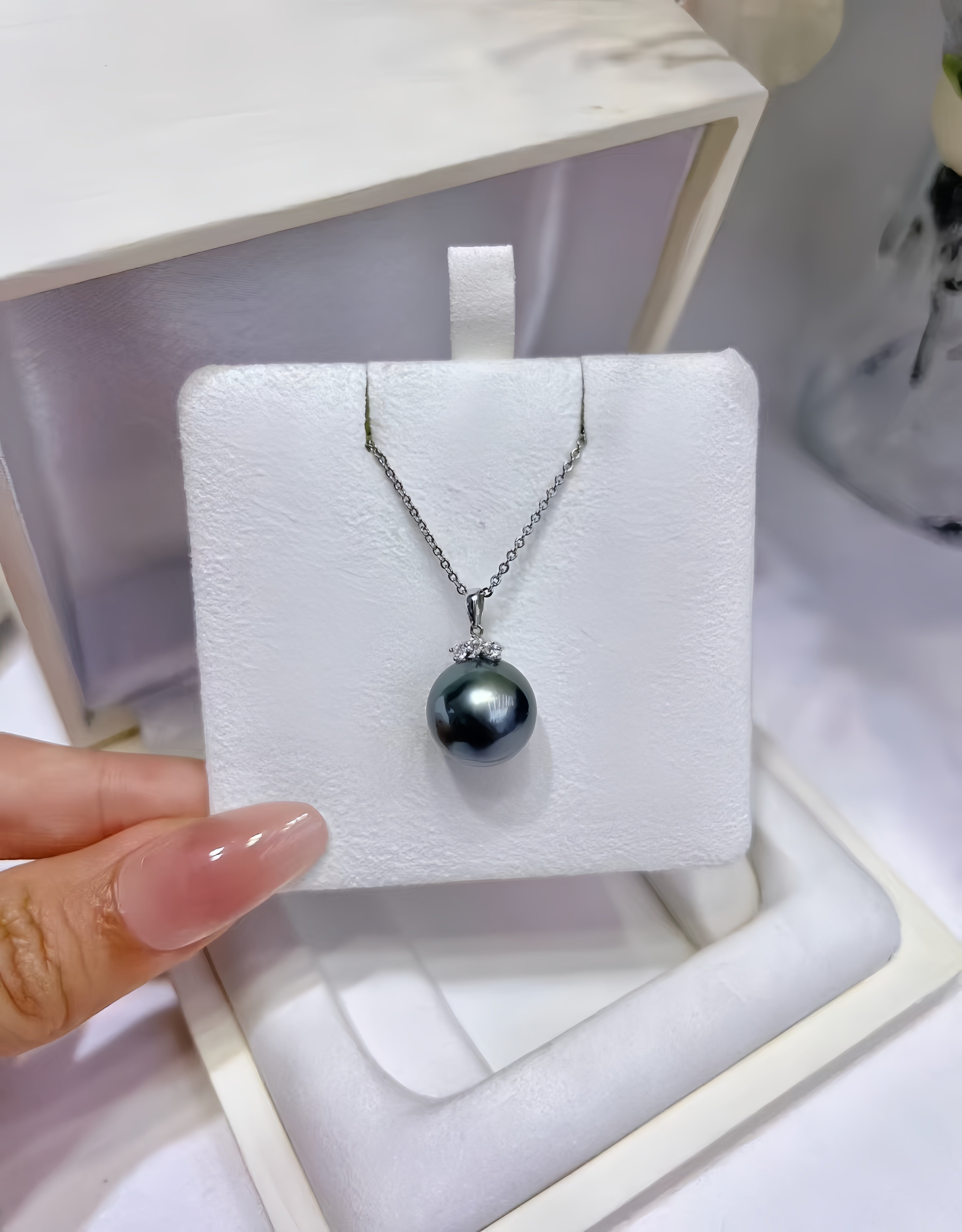 A silver chain necklace with a single dark gray pearl pendant displayed on a white jewelry stand, held by a hand with manicured nails.