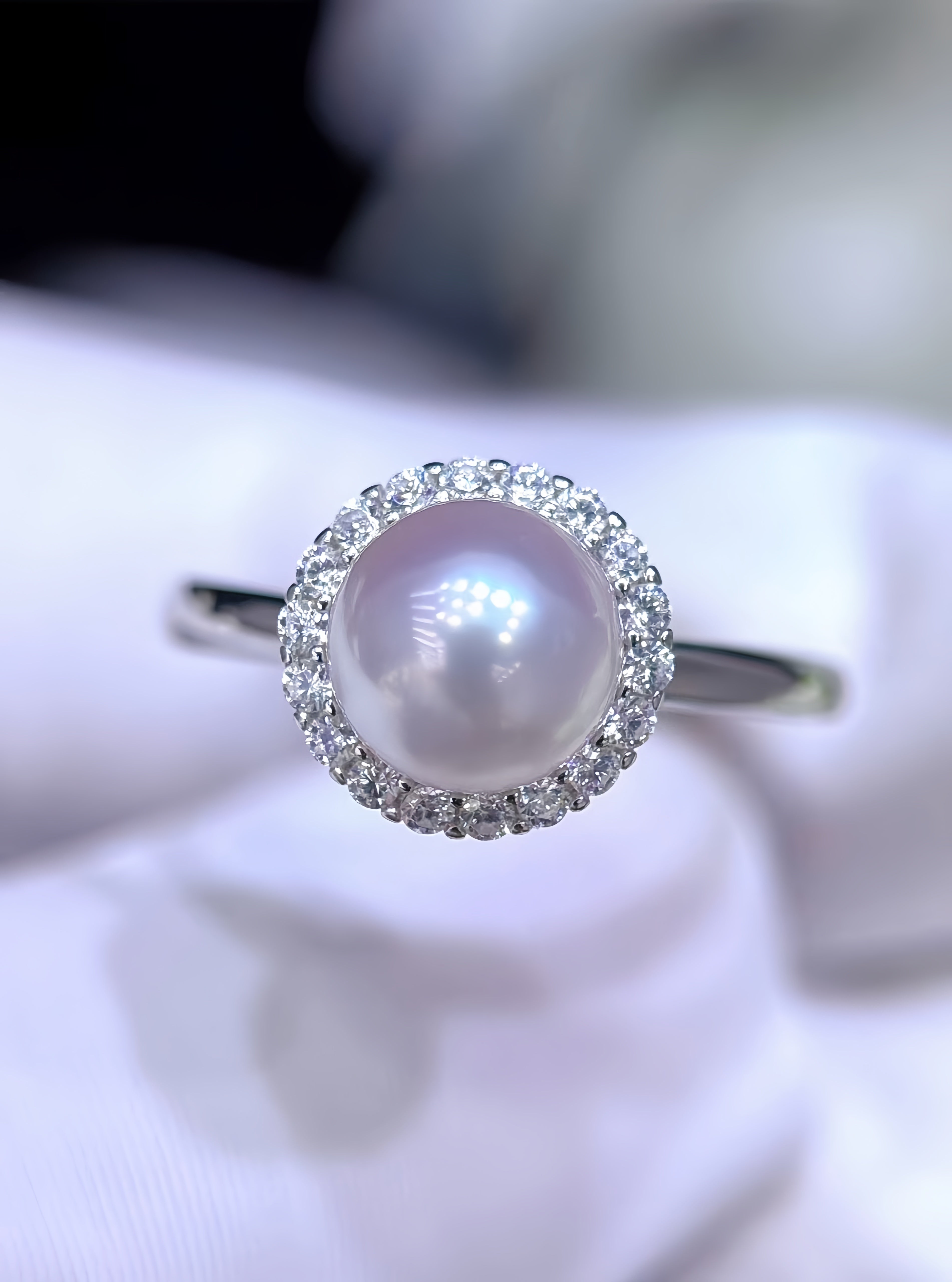 A silver ring featuring a round lustrous pearl surrounded by a halo of small clear stones, displayed on a soft white fabric surface.