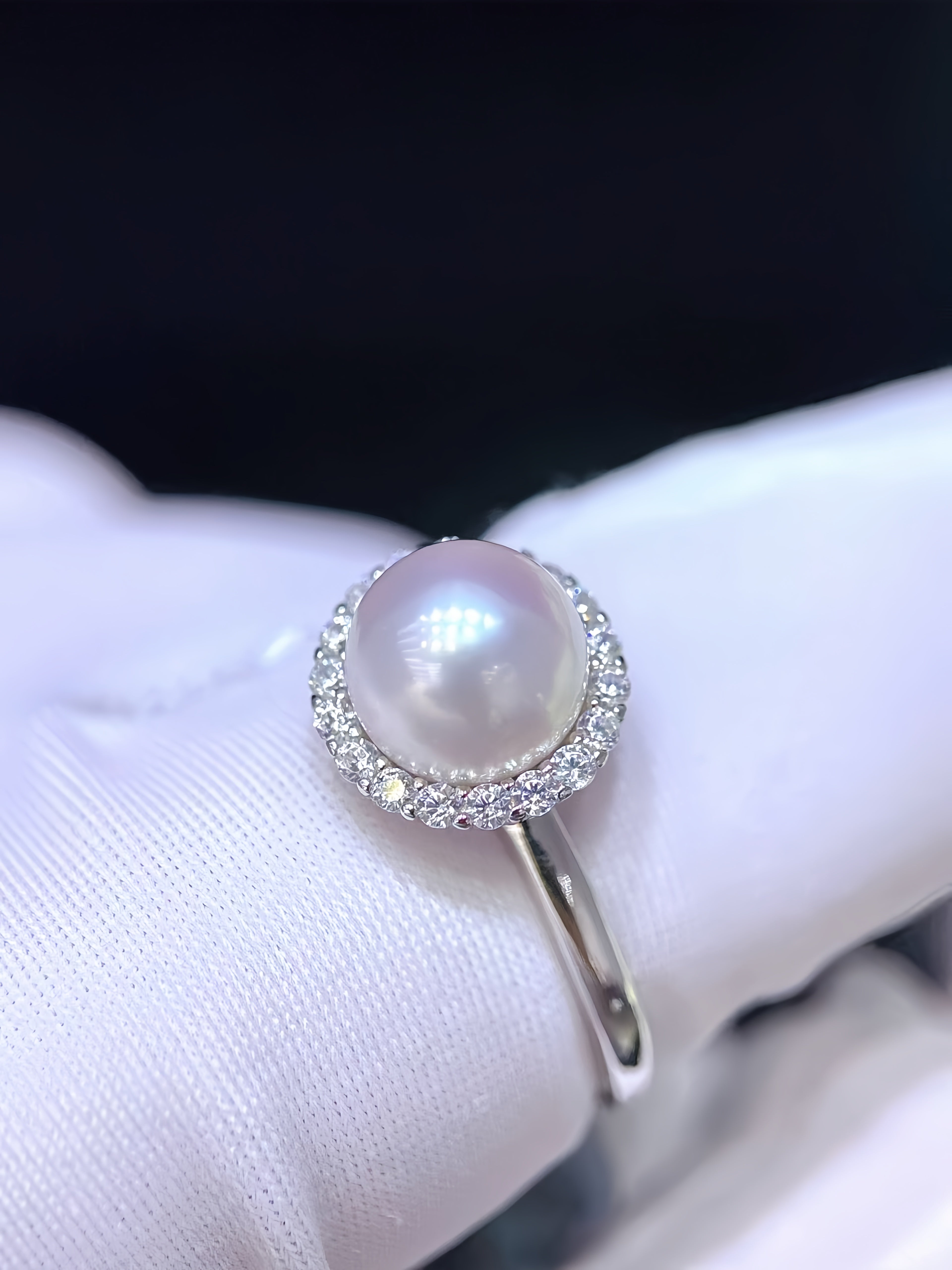 A silver ring featuring a large round white pearl surrounded by a halo of small clear stones, shown resting on a white textured surface.