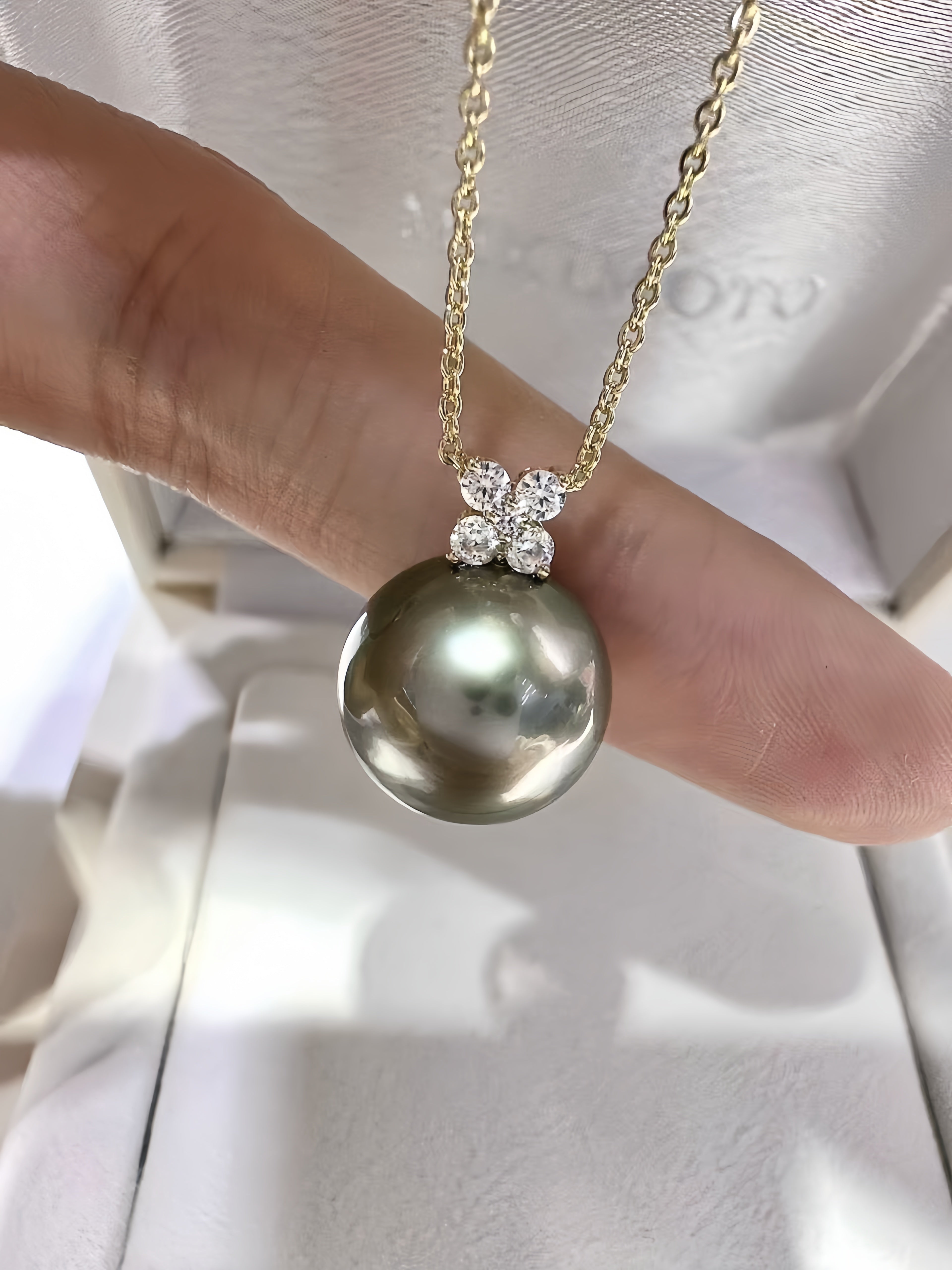 A close-up of a gold chain necklace with a large, round grayish-green pearl pendant, held by a person's fingers against a white jewelry box background.