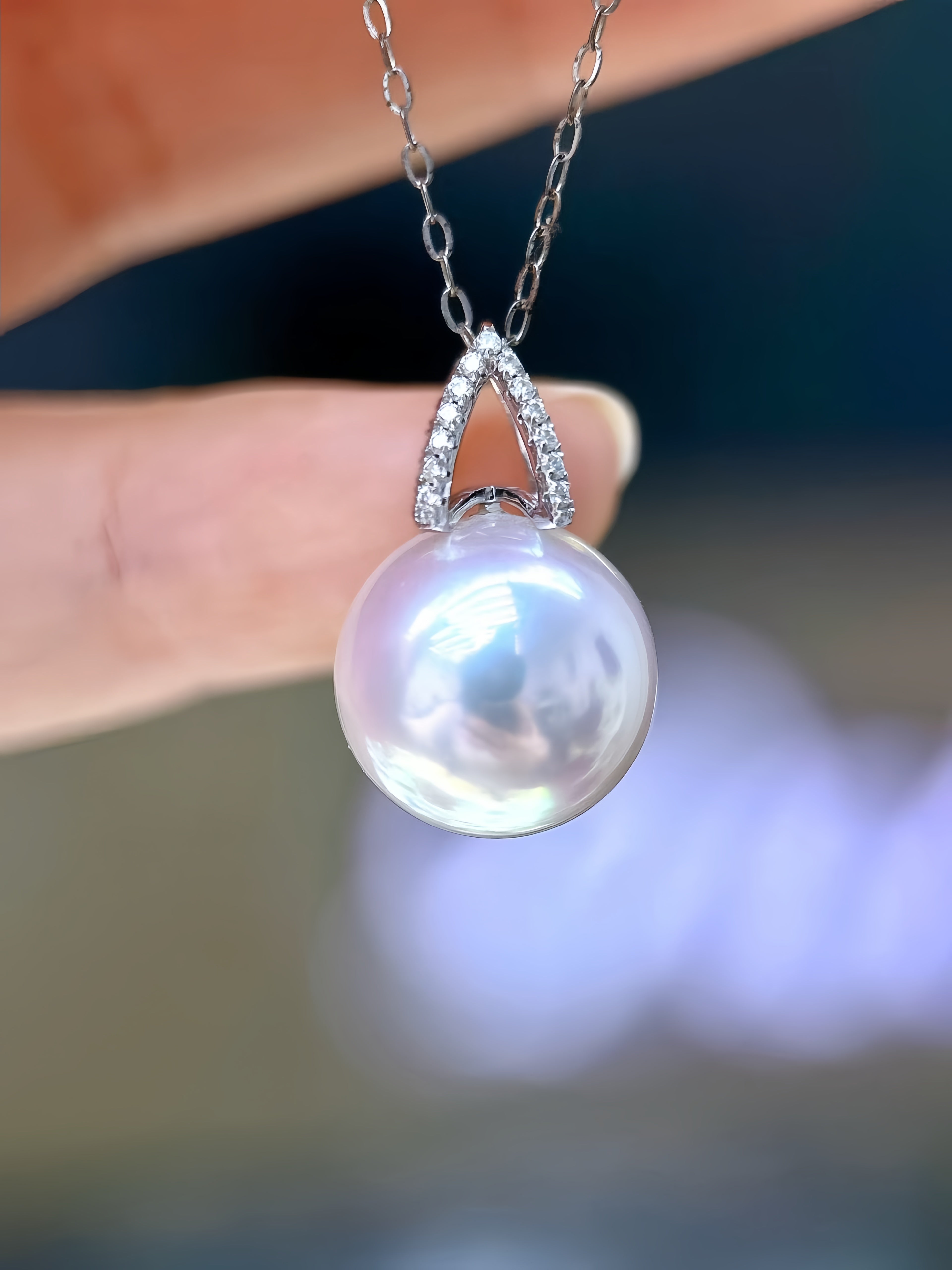 A close-up of a silver pendant necklace with a large, lustrous white pearl and a diamond-accented V-shaped top, held by fingers against a blurred background.