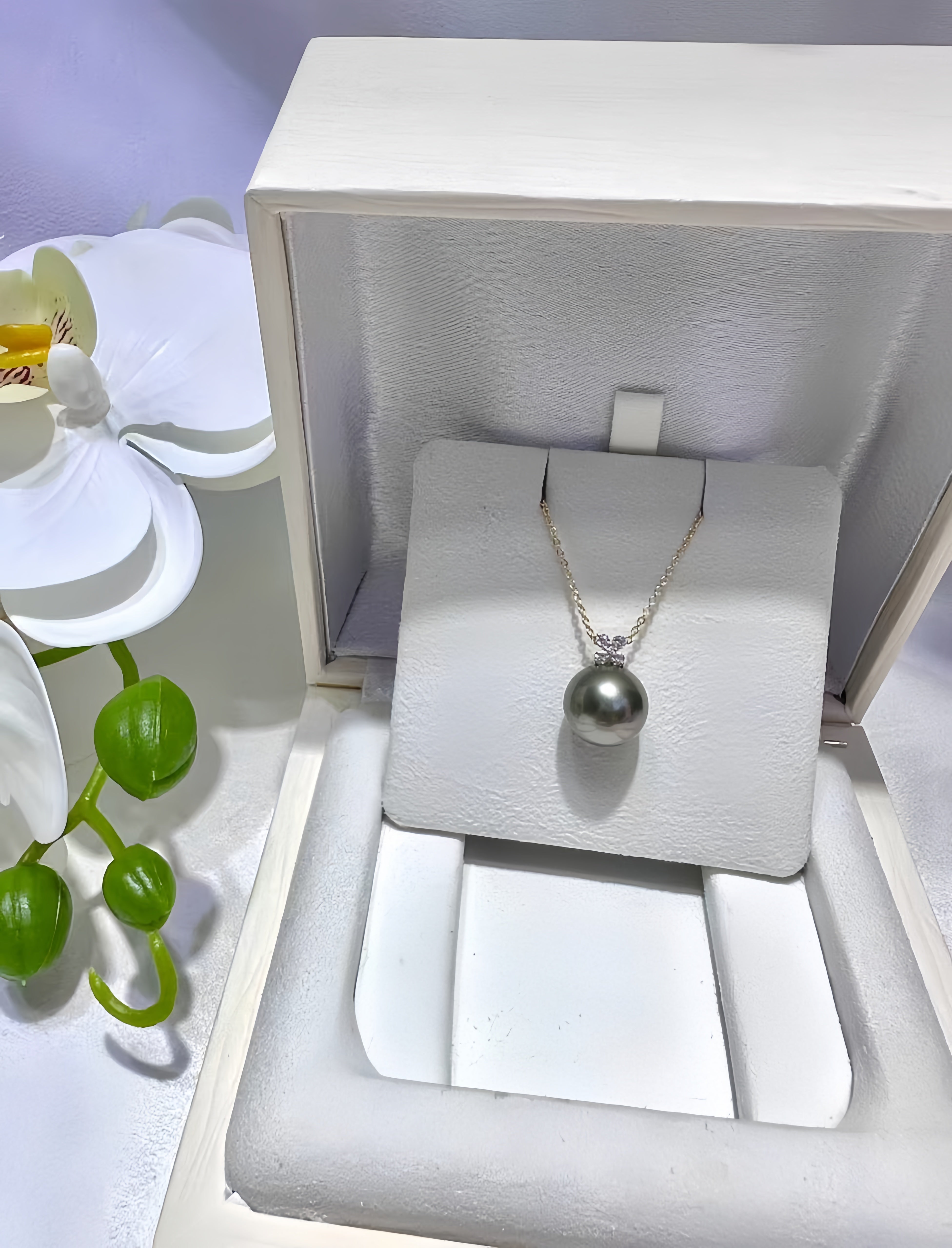 A silver necklace with a single dark gray pearl pendant displayed inside an open white jewelry box, next to a white orchid flower.