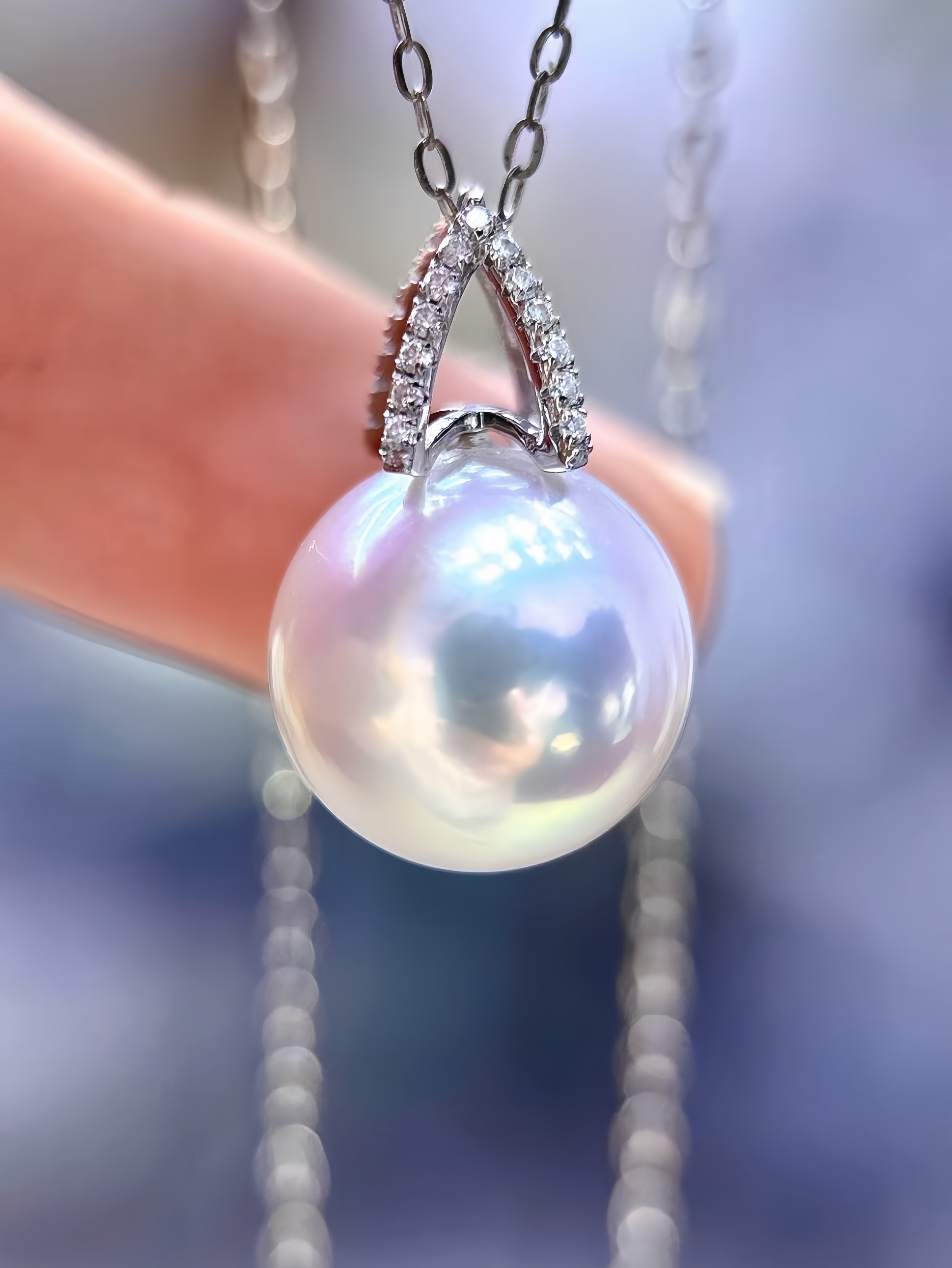 A close-up of a pearl pendant necklace held by a finger. The pendant features a large, lustrous white pearl suspended from a silver-toned V-shaped setting adorned with small clear stones, with a delicate chain hanging above.