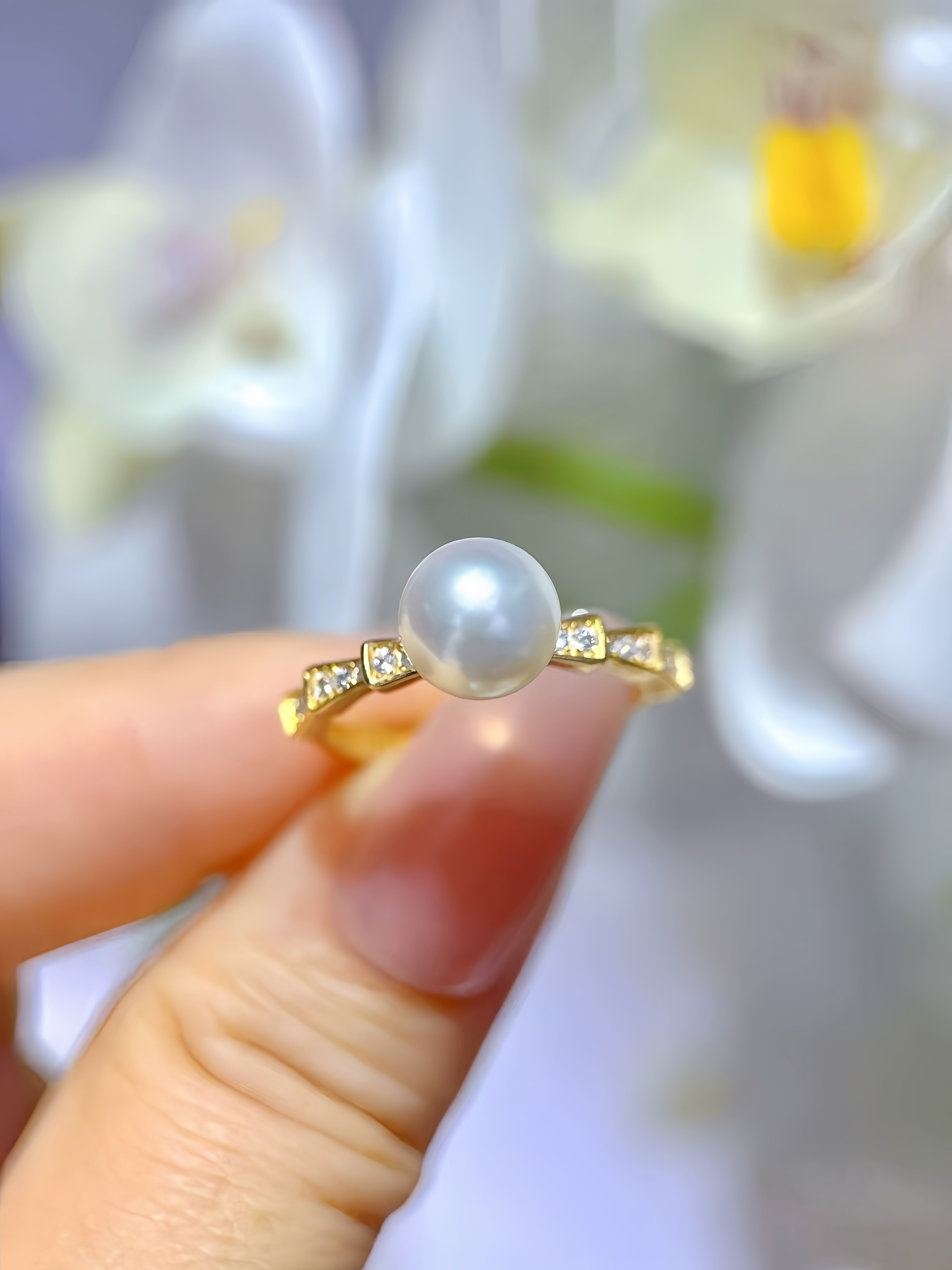 A gold ring with a single round white pearl and small clear stones on either side, held between fingers against a soft-focus background of white flowers.