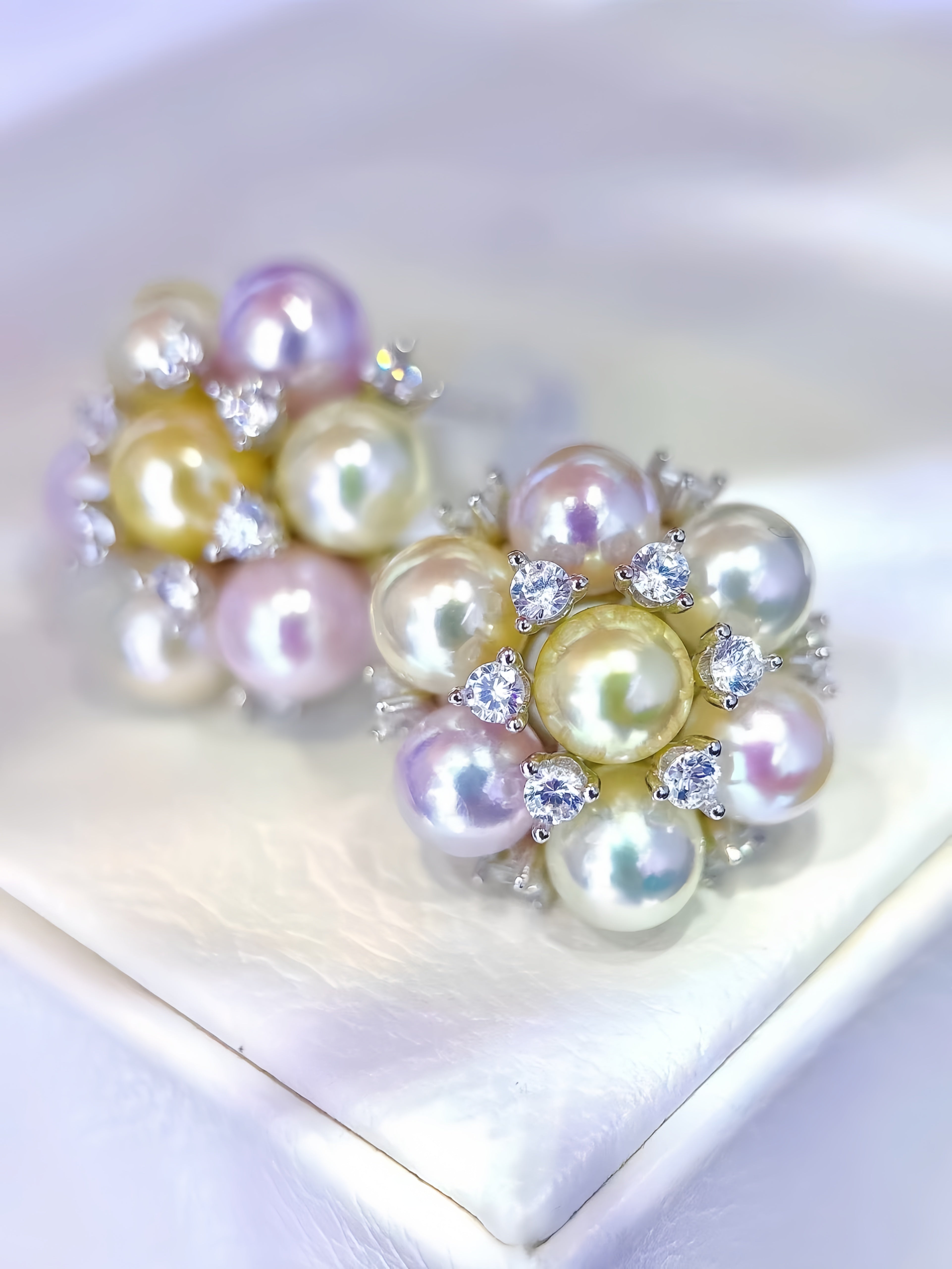 A pair of floral cluster earrings made of iridescent freshwater pearls in white, pale yellow, and lavender, accented with small clear cubic zirconia stones in silver settings, displayed on a white satin surface.