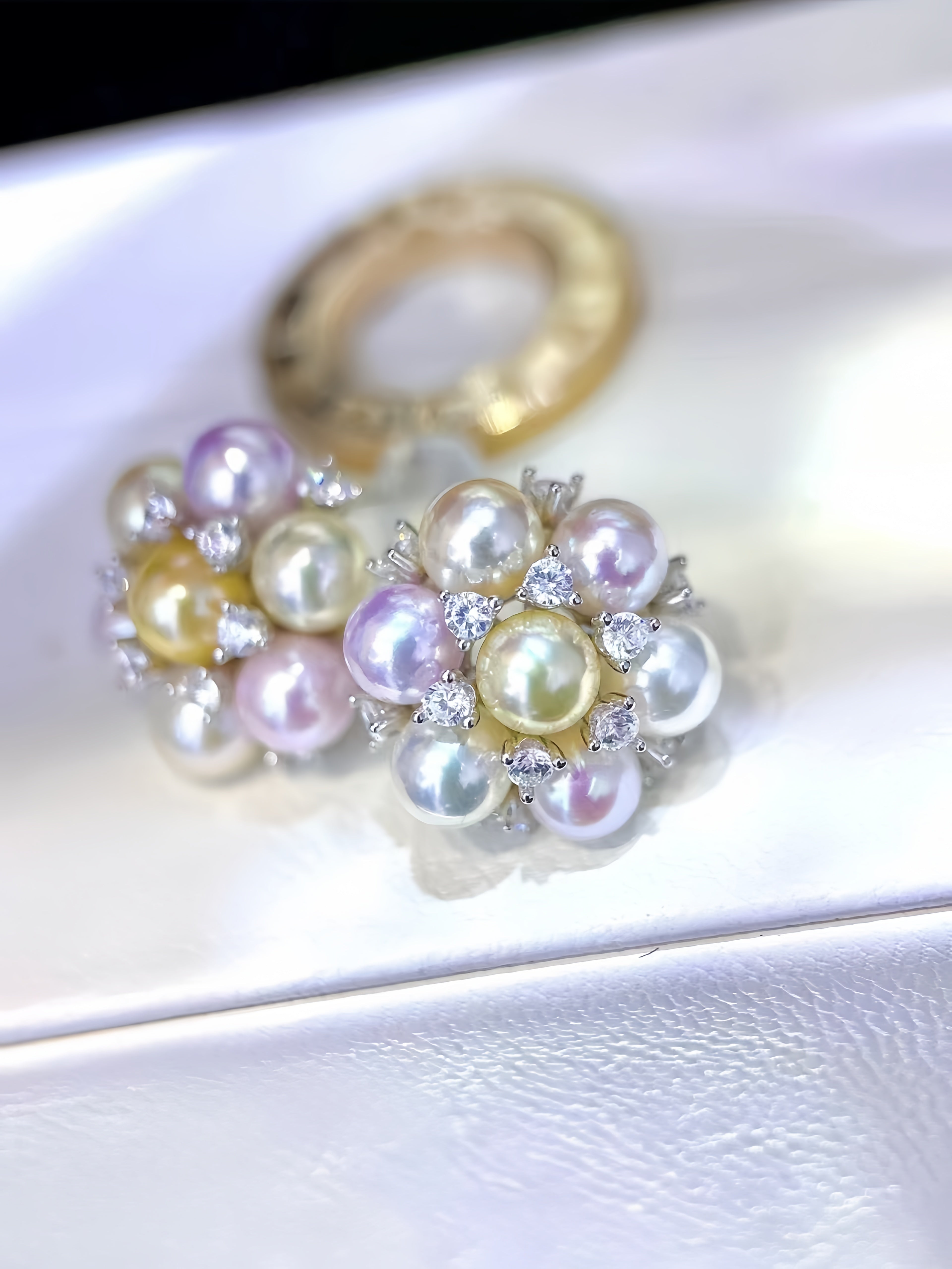 A pair of pearl flower-shaped earrings with yellow and white pearls, accented with clear rhinestones and a gold band, displayed on a white textured surface.