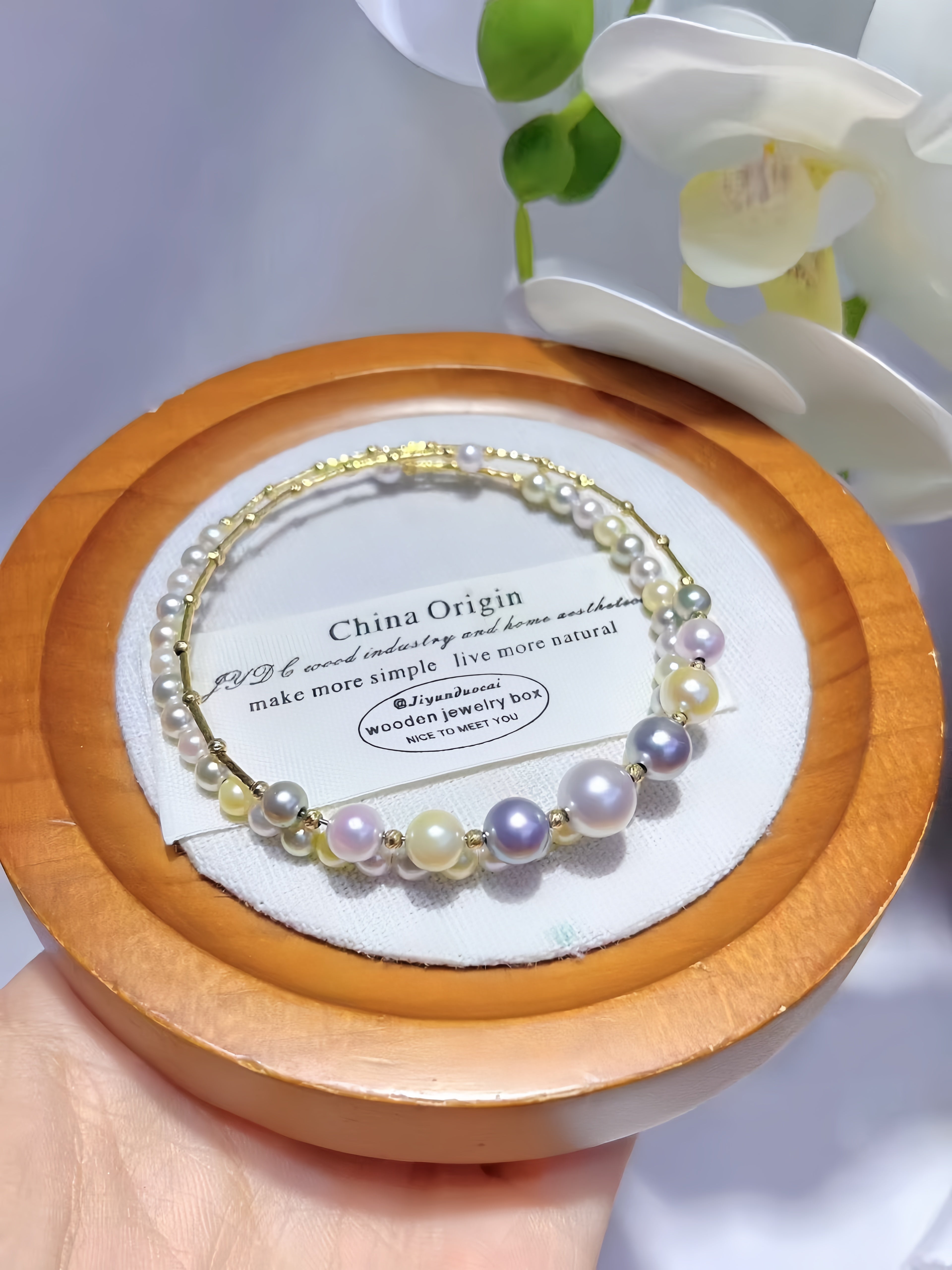 A delicate bracelet with white, lavender, and yellow pearls on a gold-tone chain, displayed on a white card inside a round wooden box held by a hand.