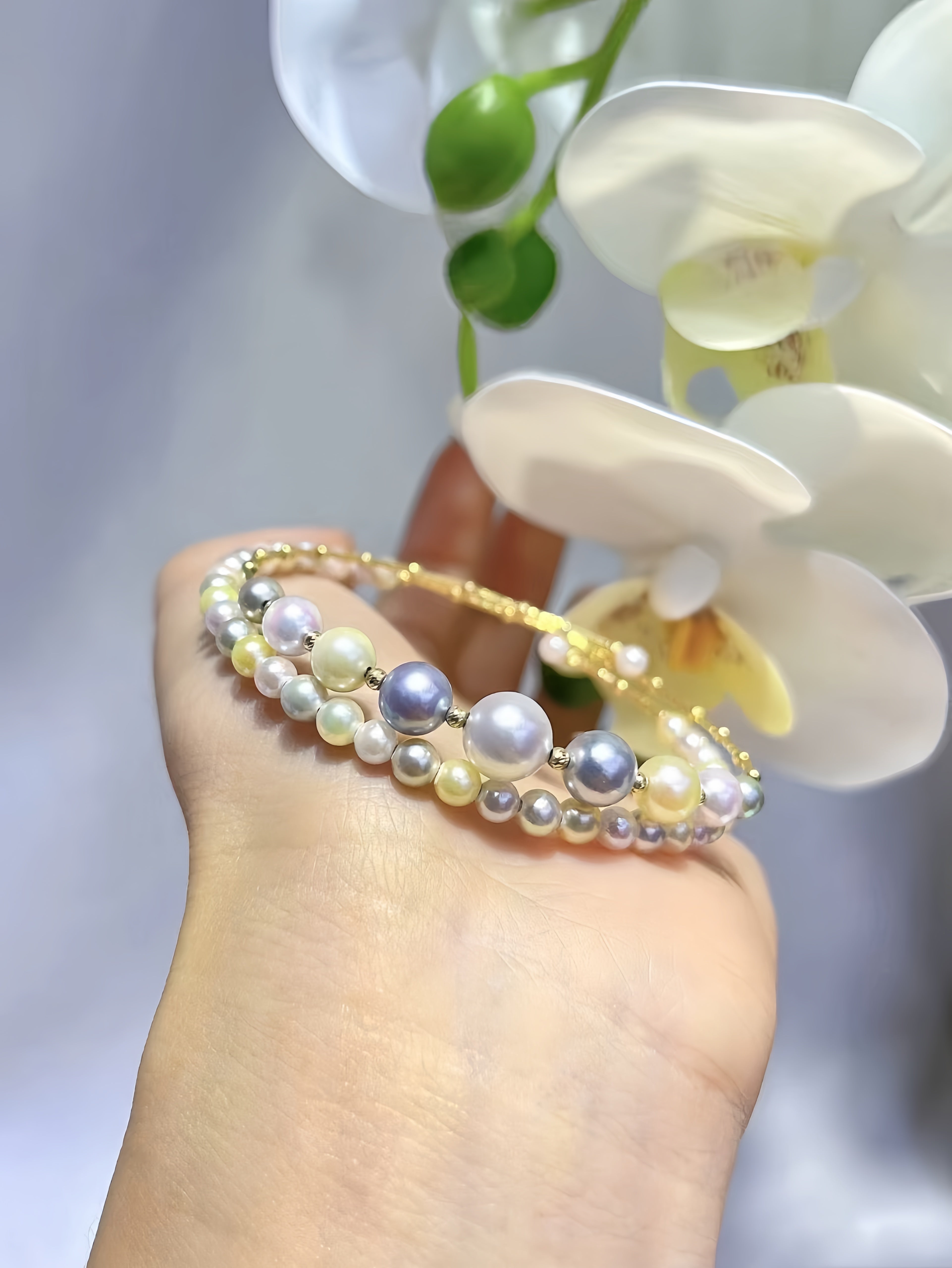 A close-up of a hand wearing a delicate double-strand beaded bracelet with white, gray, and lavender pearls on a gold-tone band, with white orchid flowers in the background.
