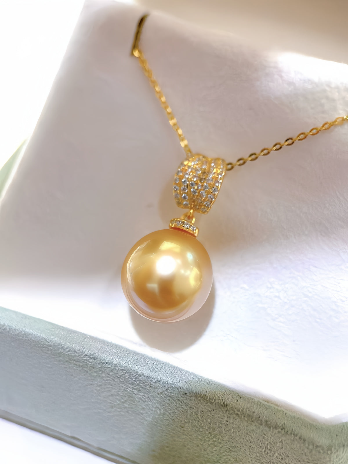 A gold chain necklace with a large round golden pearl pendant and a sparkling gold bead detail, displayed on a white jewelry box.