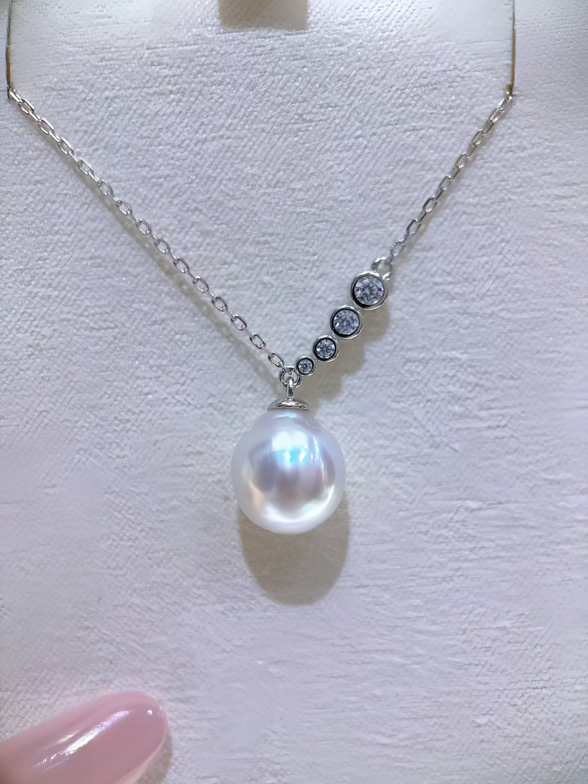 A silver necklace with a large white freshwater pearl pendant and four small round clear stones along the chain, displayed on a textured white jewelry bust.