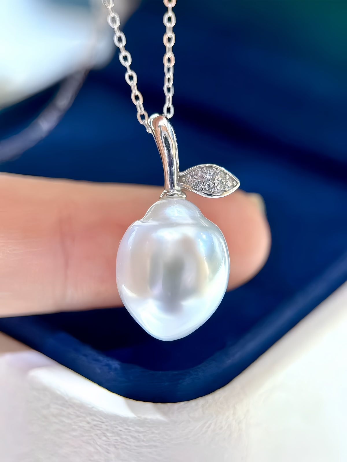 A close-up of a silver pendant necklace shaped like a pear or lemon, featuring a large lustrous white pearl and a small leaf detail with clear stones, held by a finger against a dark blue background.