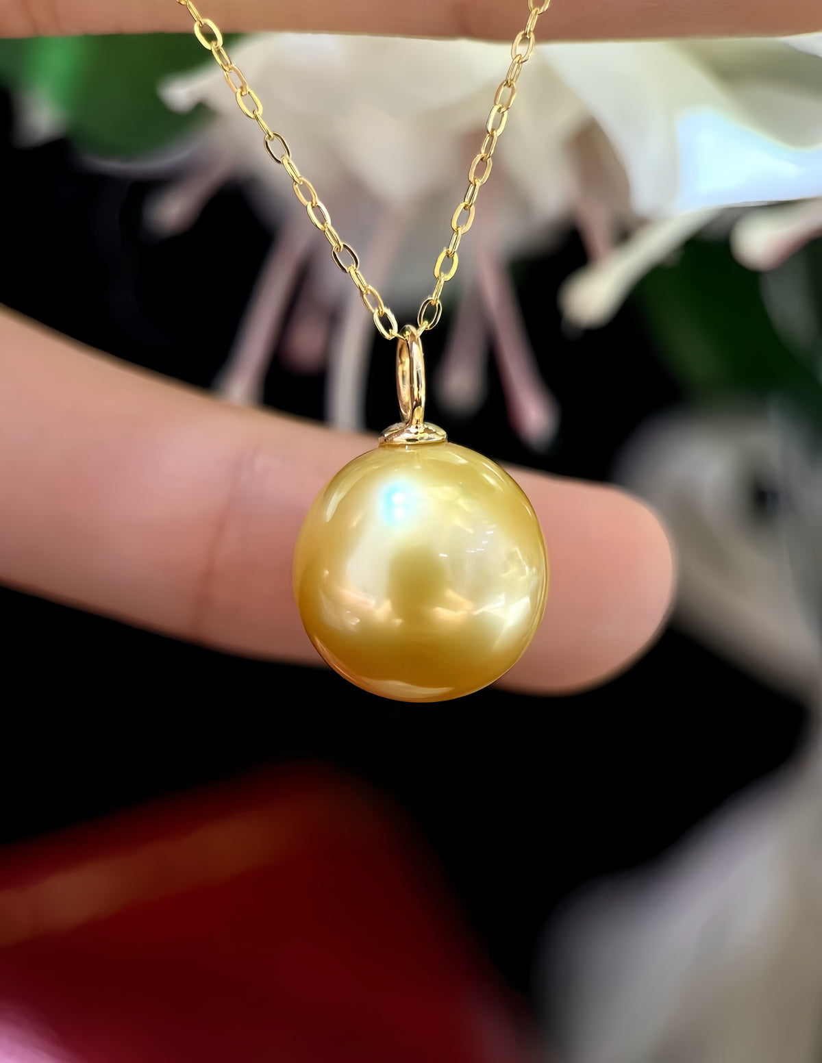 A gold pendant necklace featuring a large, round, lustrous gold pearl, held by a person's fingers against a dark, blurred background with soft white and green highlights.