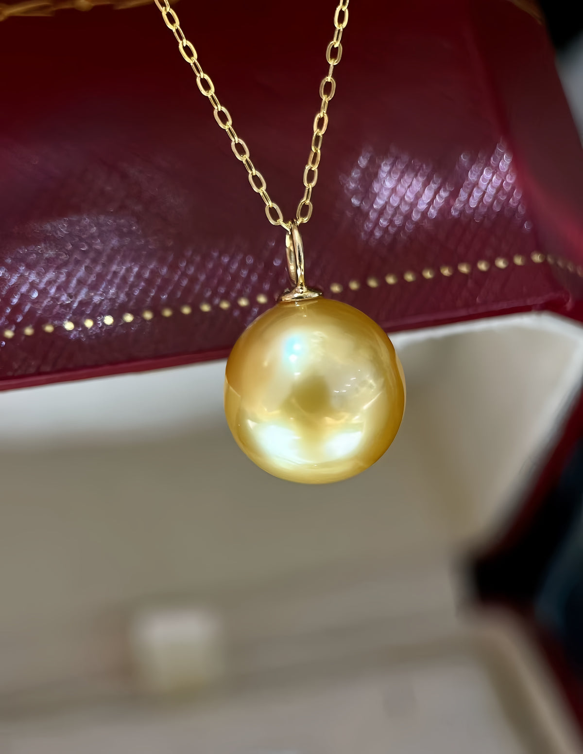 A gold chain necklace with a large, lustrous golden pearl pendant, shown hanging in front of a dark red textured box.