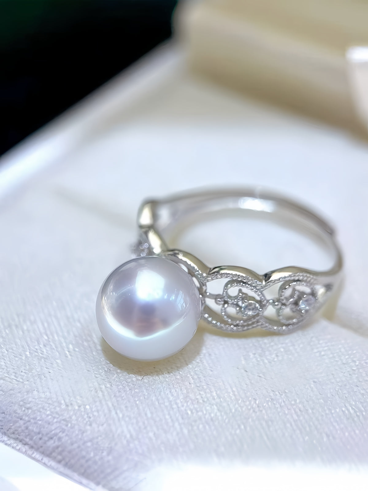 A silver ring featuring a large, lustrous white freshwater pearl and delicate filigree detailing, displayed on a soft white fabric surface.