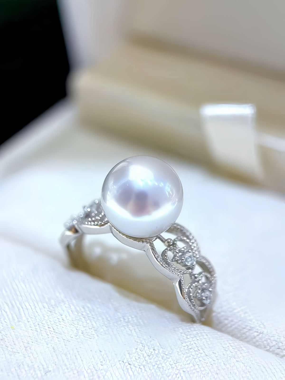 A silver ring featuring a large, lustrous white pearl in a decorative setting with small clear stones, displayed on a white textured surface with a blurred cream-colored jewelry box in the background.