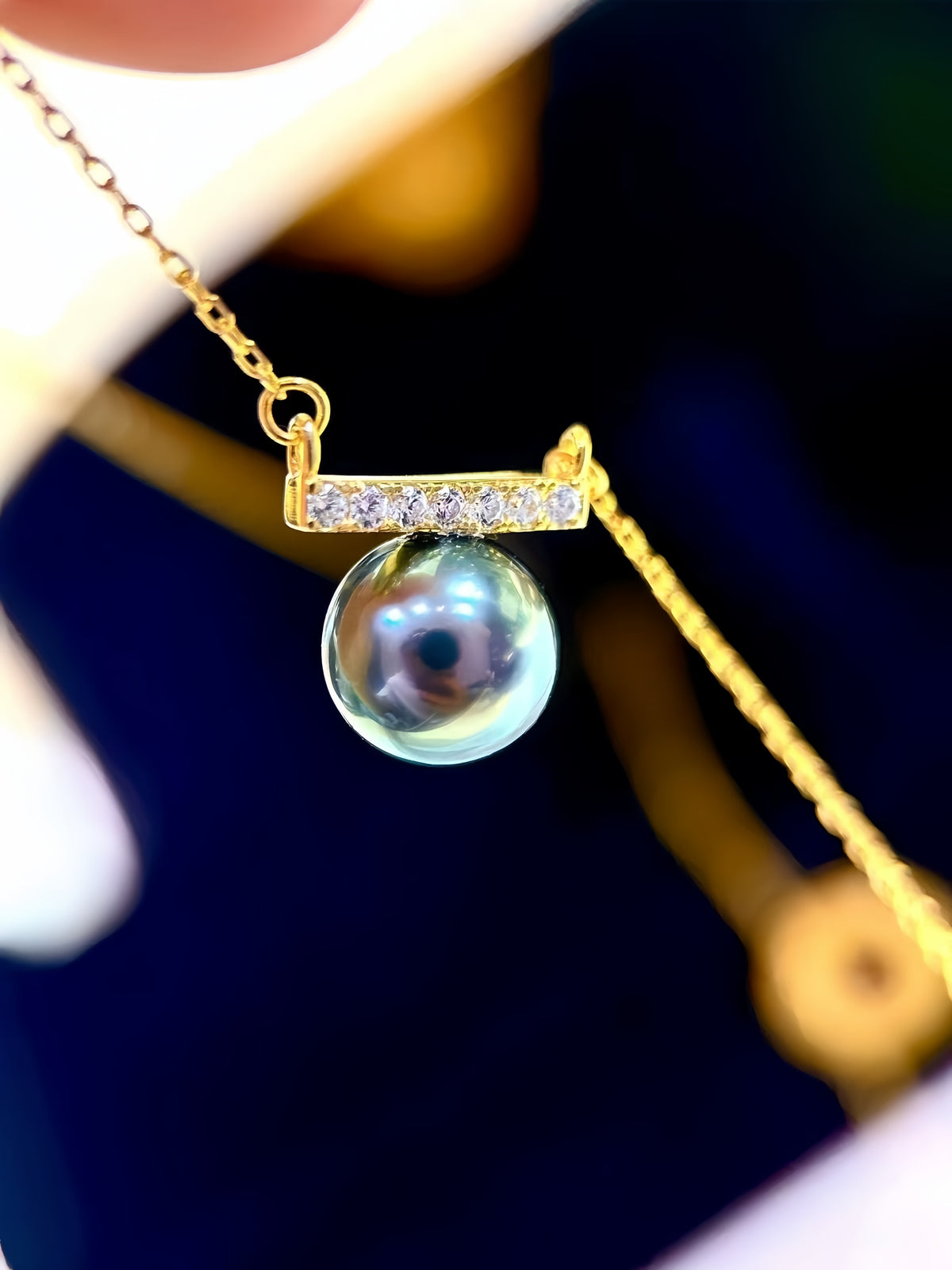 A gold pendant necklace featuring a round iridescent pearl suspended beneath a small gold bar set with clear stones, shown close up against a dark blue and white background.