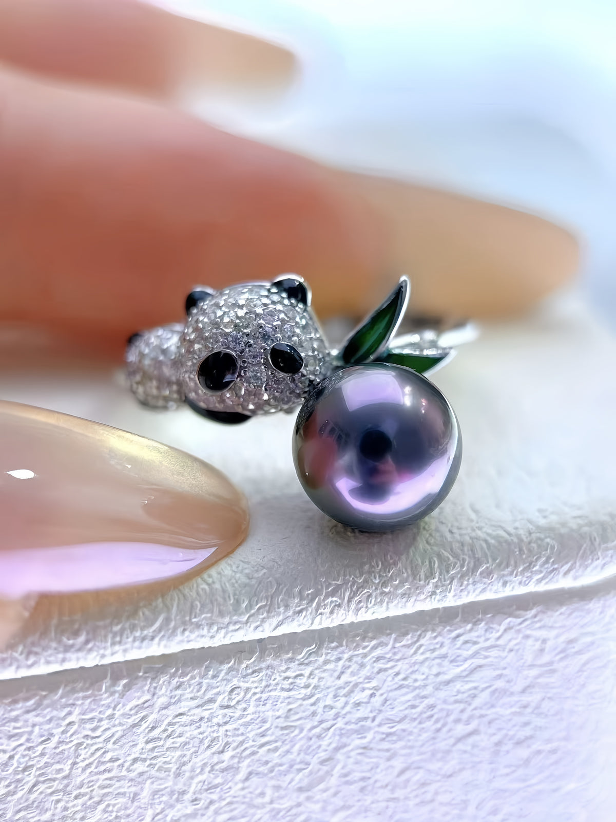 A close-up of a silver ring shaped like a panda holding a dark purple pearl fruit, resting on a person's finger against a white textured background.