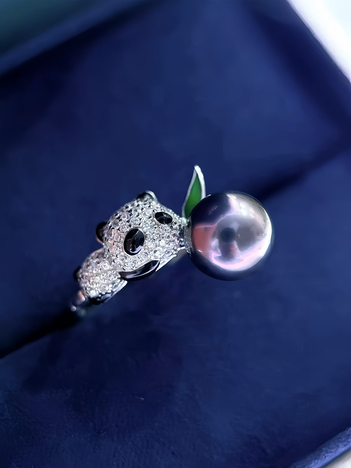 A close-up of a ring featuring a silver-toned panda face holding a large, lustrous dark pearl with a small green leaf accent, resting on a dark navy velvet ring box.