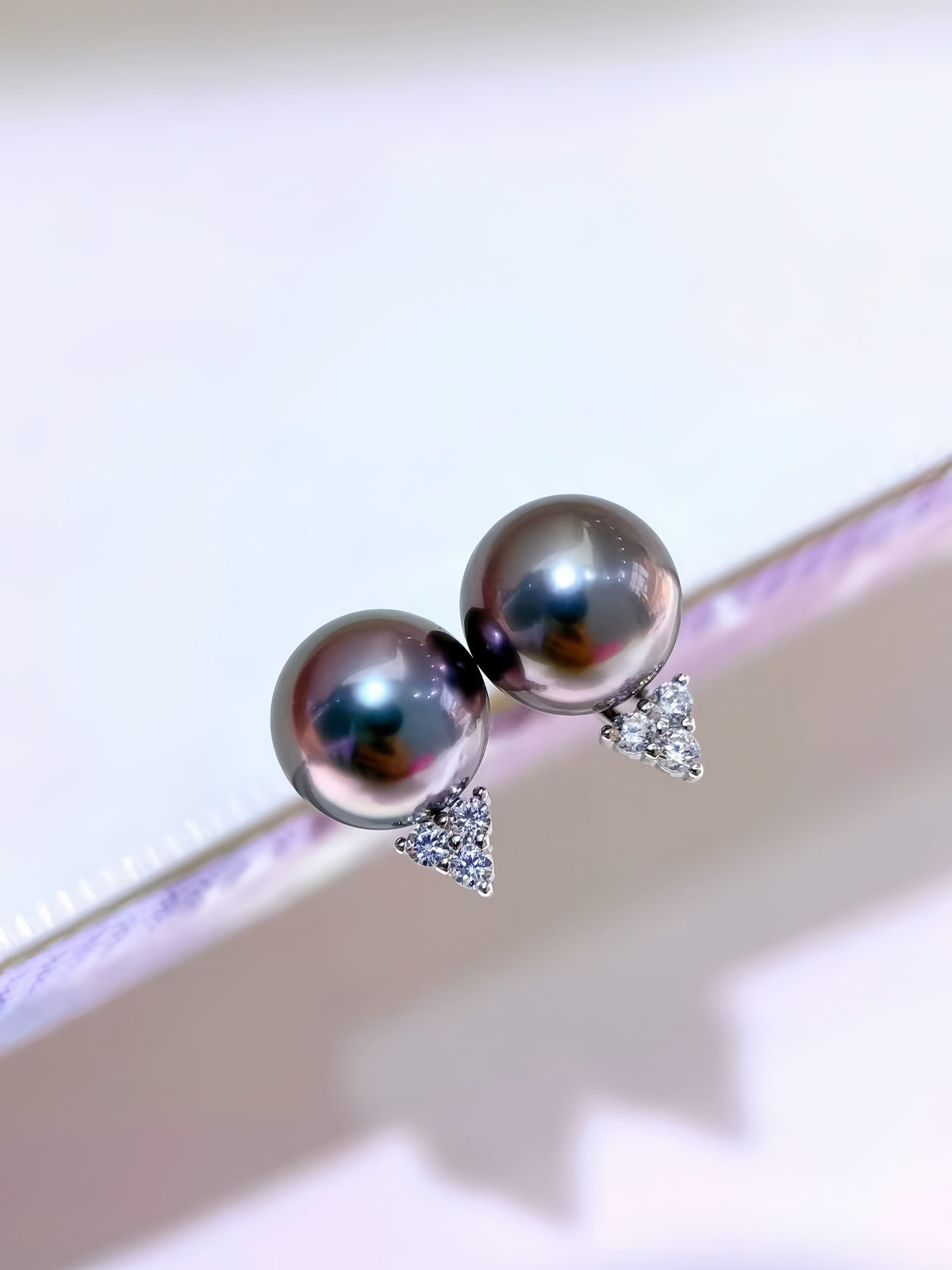 A pair of stud earrings with round dark gray pearl-like beads and small silver triangular crystal clusters, shown resting on a person's fingers against a bright white background.