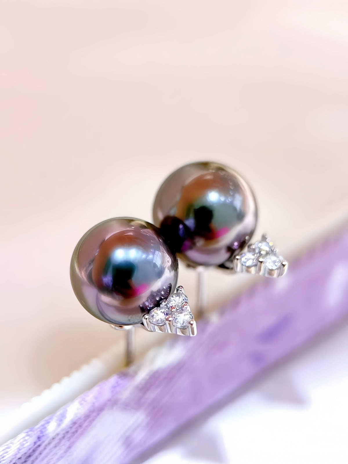 A pair of stud earrings with round dark gray pearl-like beads and small silver triangular crystal clusters, shown resting on a person's fingers against a bright white background.