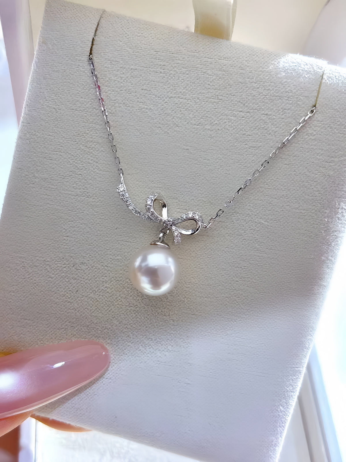 A silver chain necklace with a bow-shaped pendant set with small clear stones and a single white pearl drop, displayed on a white jewelry box held by a hand with pink nail polish.