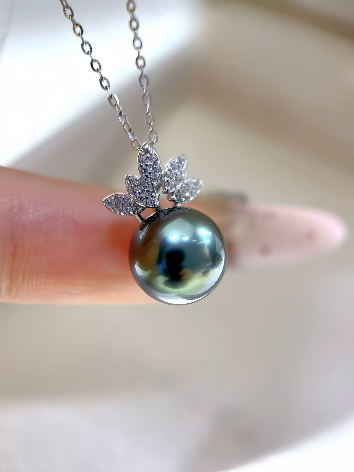 A close-up of a silver pendant necklace featuring a dark green-gray pearl apple with a silver leaf crown set with small clear stones, held by a finger against a soft beige background.