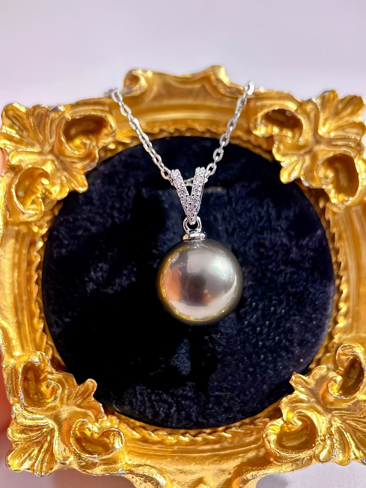 A silver pendant necklace featuring a large, lustrous pearl pendant, displayed on a black velvet cushion inside an ornate gold frame.