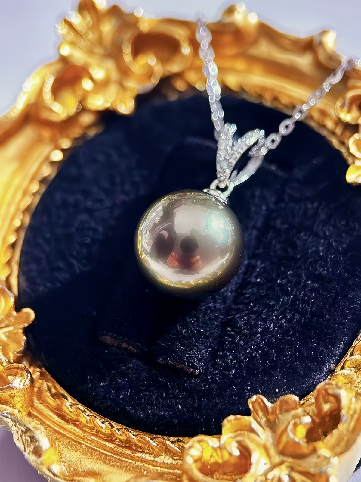 A silver pendant necklace with a round iridescent pearl resting on a black velvet cushion inside an ornate gold frame.