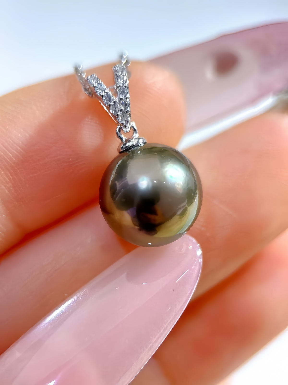 A close-up of a sterling silver pendant necklace featuring a single round, dark olive-gray Tahitian pearl, held between fingers against a soft pink and white background.